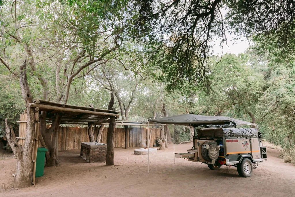 A small outdoor seating area with a makeshift wooden roof, a brick table and a wooden bench, next to a trailer camper with an extended awning, surrounded by trees in a wooded setting.