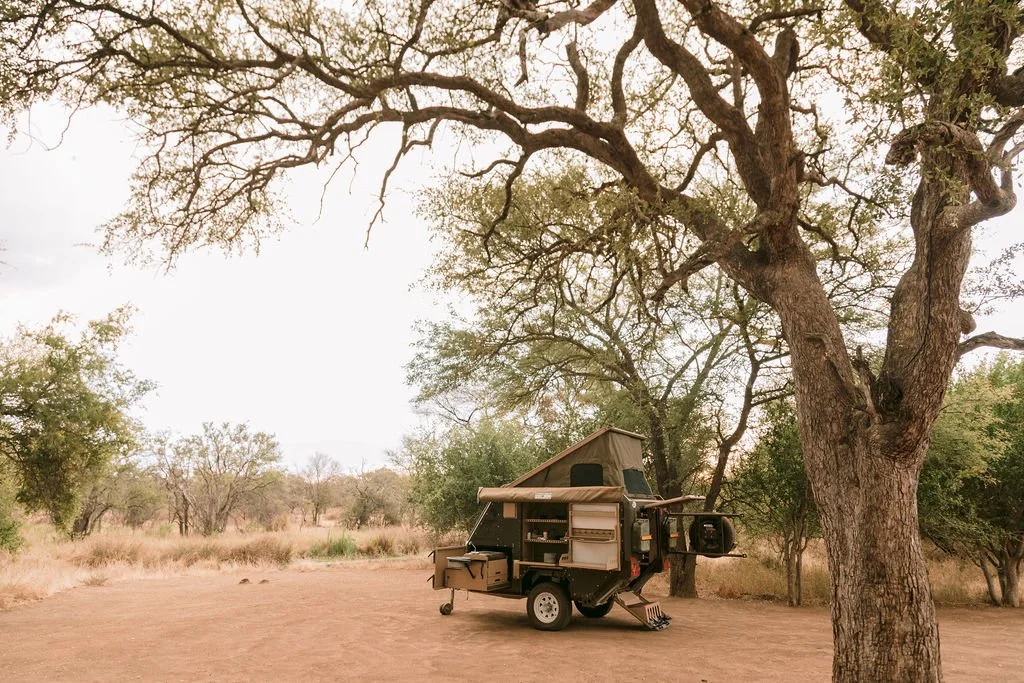 A small camping trailer with a roof tent parked under a large tree in a dry, open landscape with scattered trees and grasses.