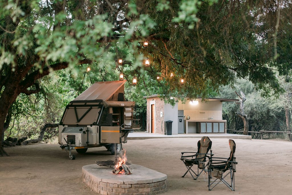 Campfire with two chairs under a large, leafy tree with hanging lanterns, a food trailer, and a small building or kitchenette in the background, set in a wooded outdoor area at dusk.