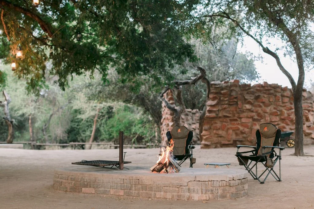 Two camping chairs and a small table around a campfire on a brick circular fire pit in an outdoor setting with trees and rocks in the background.