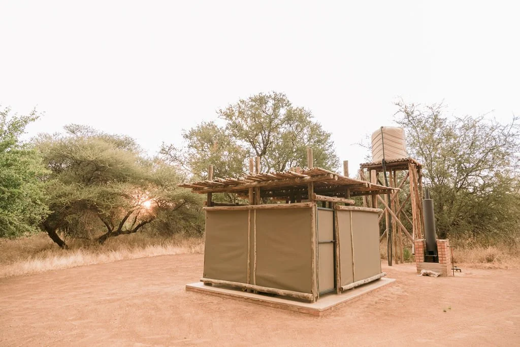 A rustic water tower and shelter structure in a dry, savannah landscape with trees and sunset in the background.