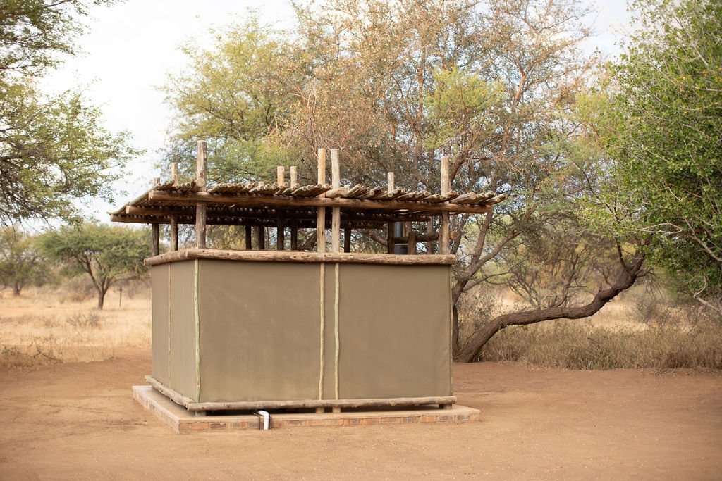 A small, rustic animal shelter made of wooden poles and cloth, situated in a dry, rural area with trees in the background.