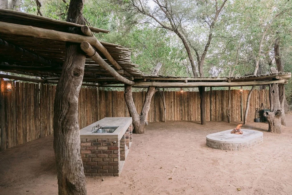Outdoor patio area with a rustic wooden fence, trees, a brick countertop with a sink, and a circular fire pit with a small fire on top.