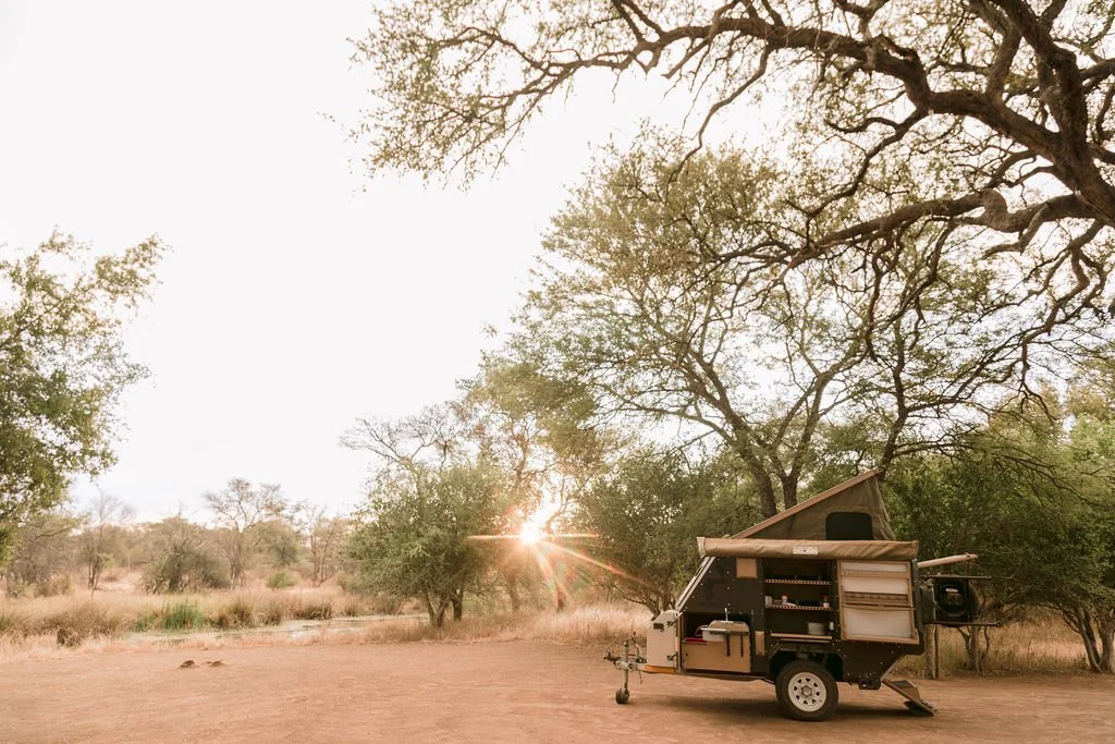 A small black mobile camper on wheels parked on dirt ground in a natural setting with trees and bushes, during sunset or sunrise.