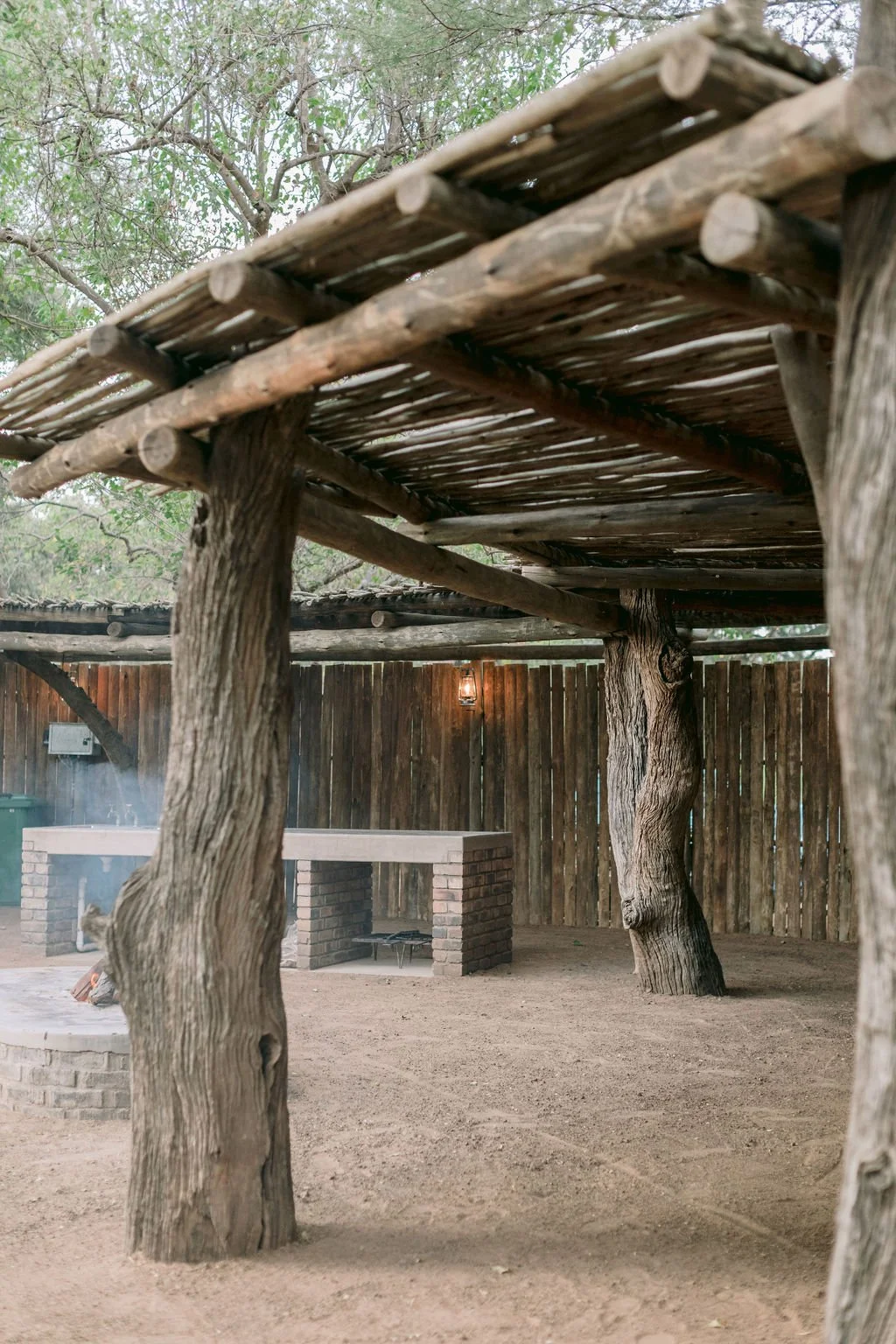 A rustic outdoor patio area with a wooden roof supported by twisted tree trunks, a brick barbecue, a wooden fence, and a lantern on a wall.