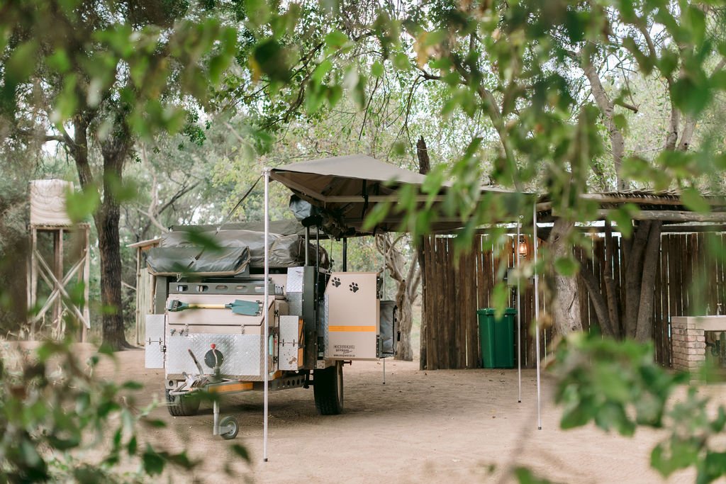 A trailer with a canopy, camping gear, and tools parked on a dirt ground, surrounded by trees and a rustic wooden fence.