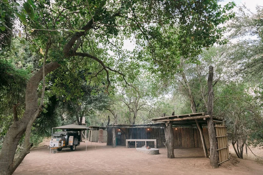 A rustic outdoor setting with a wooden structure, trees, a covered food truck, and a fire pit on a dirt surface.