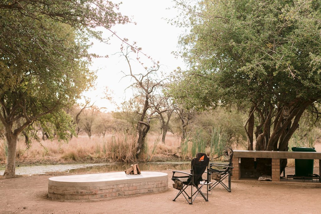 Outdoor campsite with two camping chairs, a small table, a picnic table, and a fire pit near a river, surrounded by trees and grassy area.
