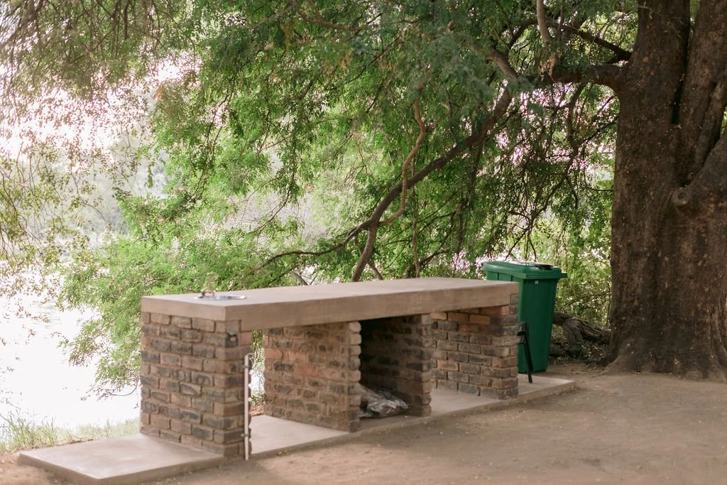 Outdoor scene with a brick picnic table, a large tree with spreading branches, and a green trash bin beside the tree on a concrete platform.