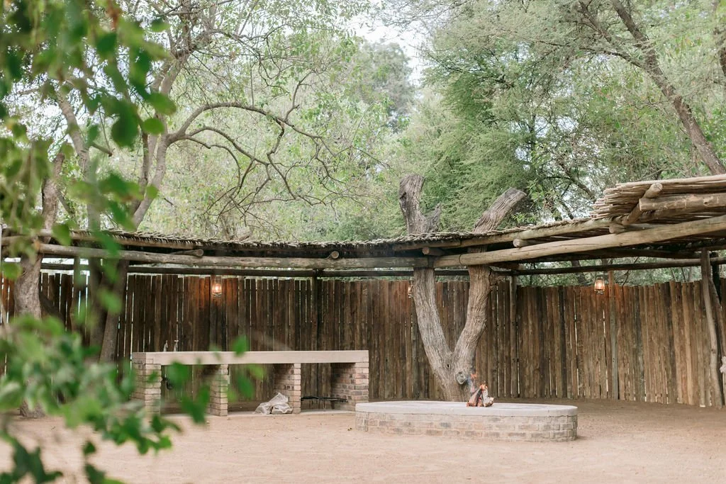 An outdoor area with a dirt ground, a wooden fence, and large trees providing shade. There is a circular brick platform with a small dog resting on it, and a rustic wooden counter or bench is visible against the fence. The area has a natural, rustic feel with minimal lighting.