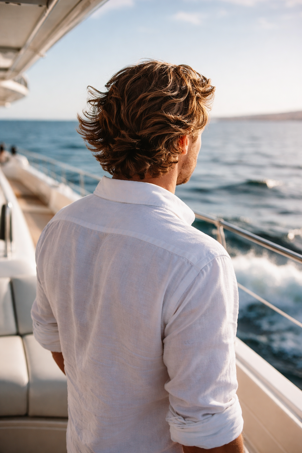 A man with wavy brown hair in a white shirt looking out at the ocean from a boat during sunset.