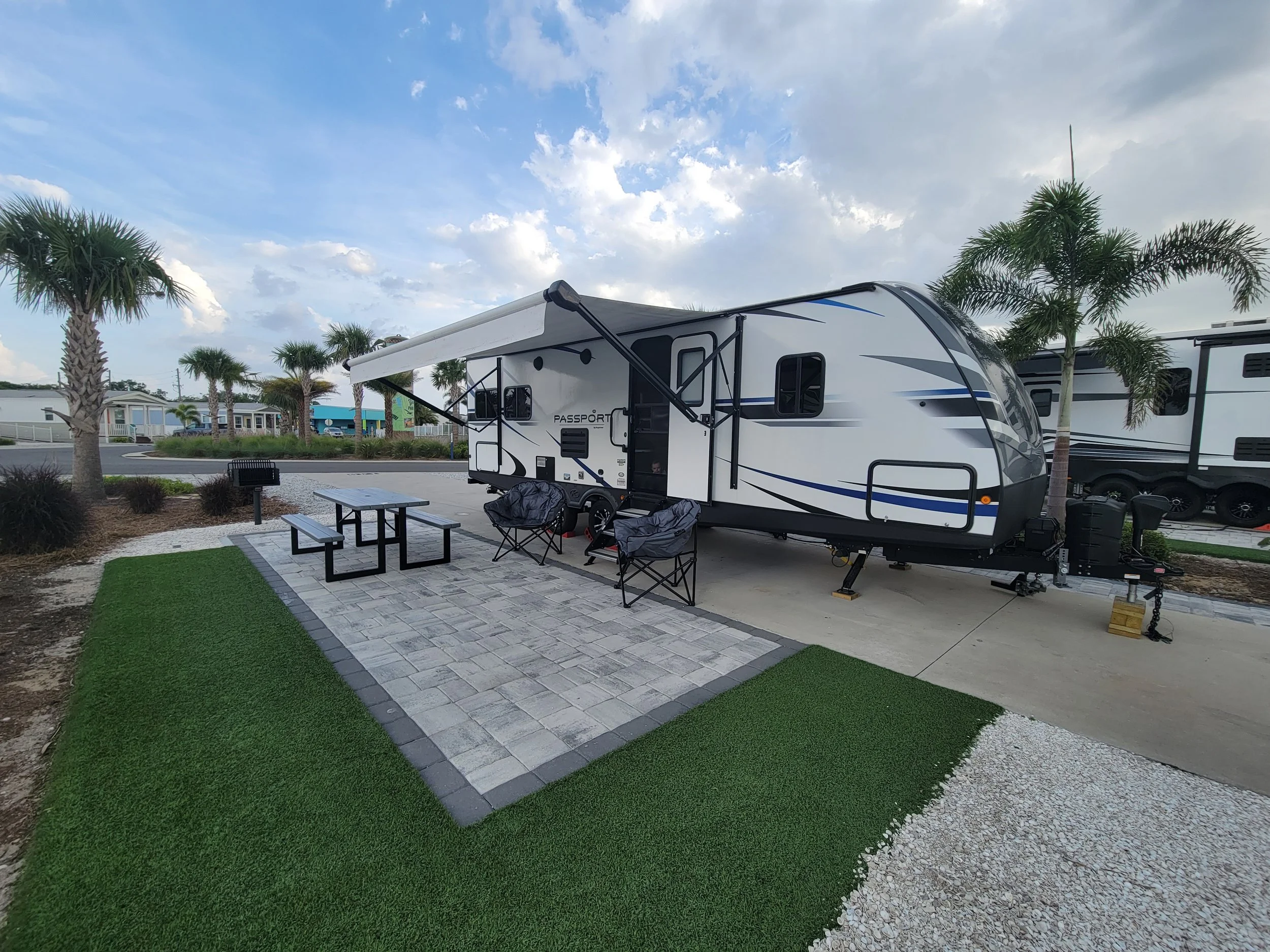 A travel trailer parked on a paved campsite with artificial grass, surrounded by palm trees, with outdoor furniture and a partly cloudy sky.