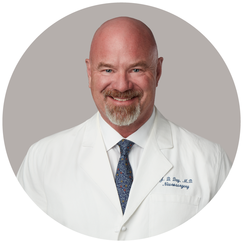 Professional portrait of a bald man with a goatee, smiling, wearing a white doctor's coat with embroidered name and specialty, and a patterned tie, against a light gray background.