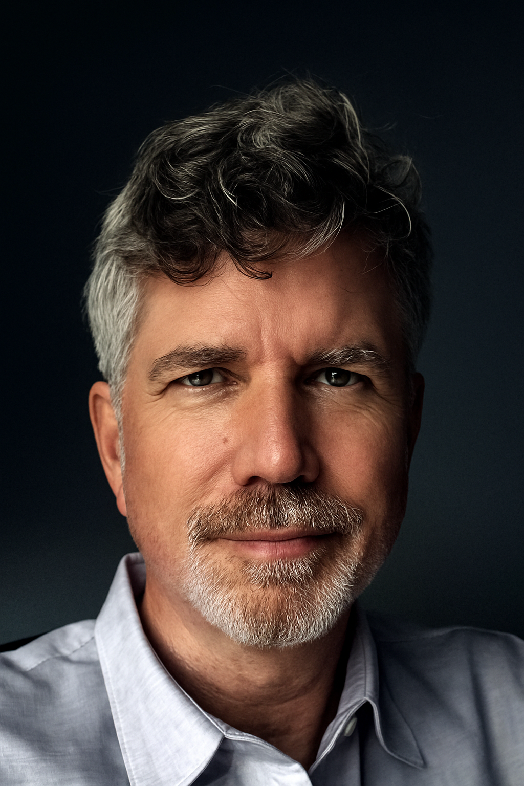 Close-up portrait of a middle-aged man with gray hair and beard, wearing a light blue collared shirt, against a dark background.