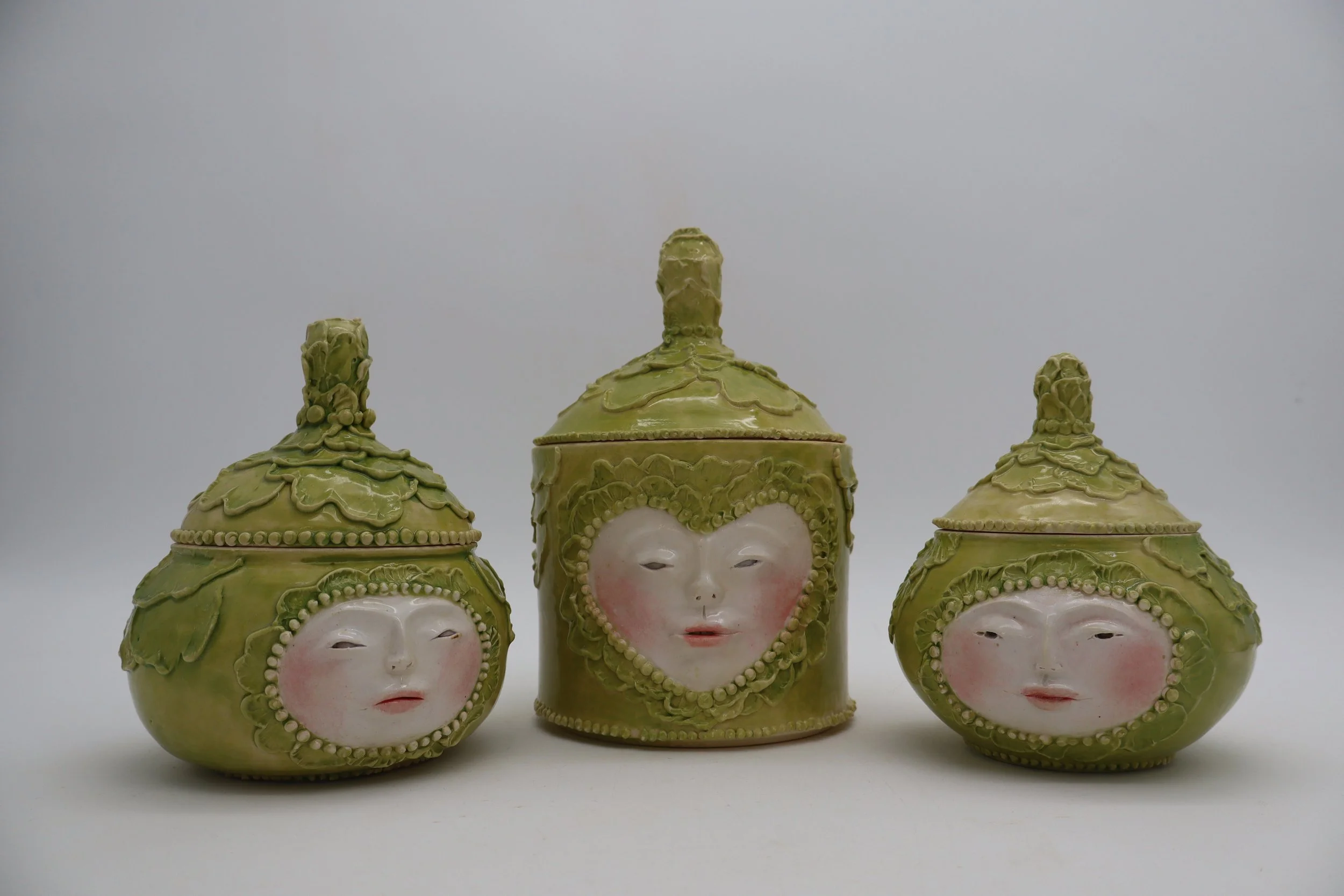 Three decorative jars with lid toppers, each featuring a porcelain face with pink cheeks, set against a plain background.