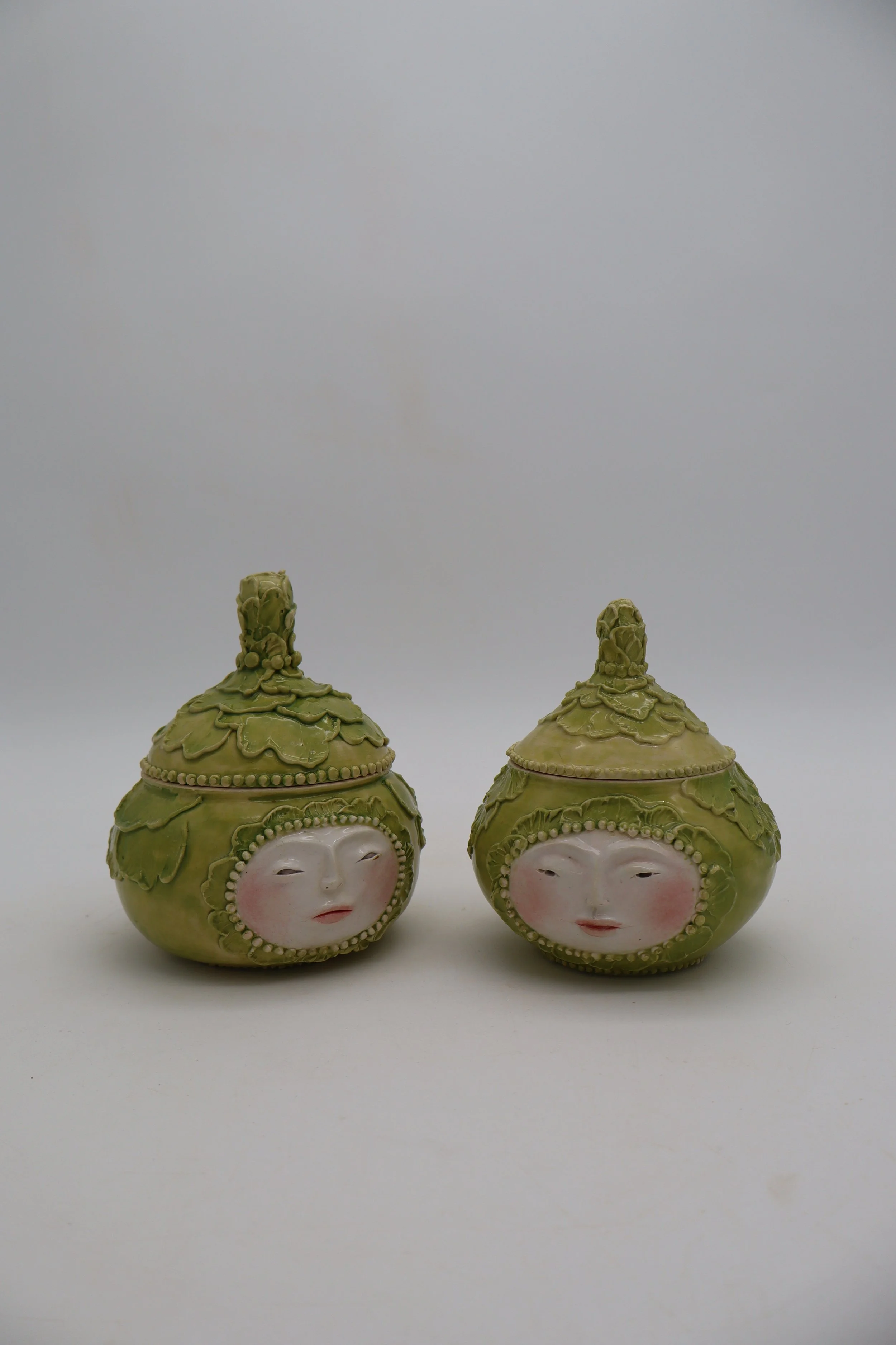 Two decorative jars with human face designs and green ornate lids, placed on a white surface against a plain white background.