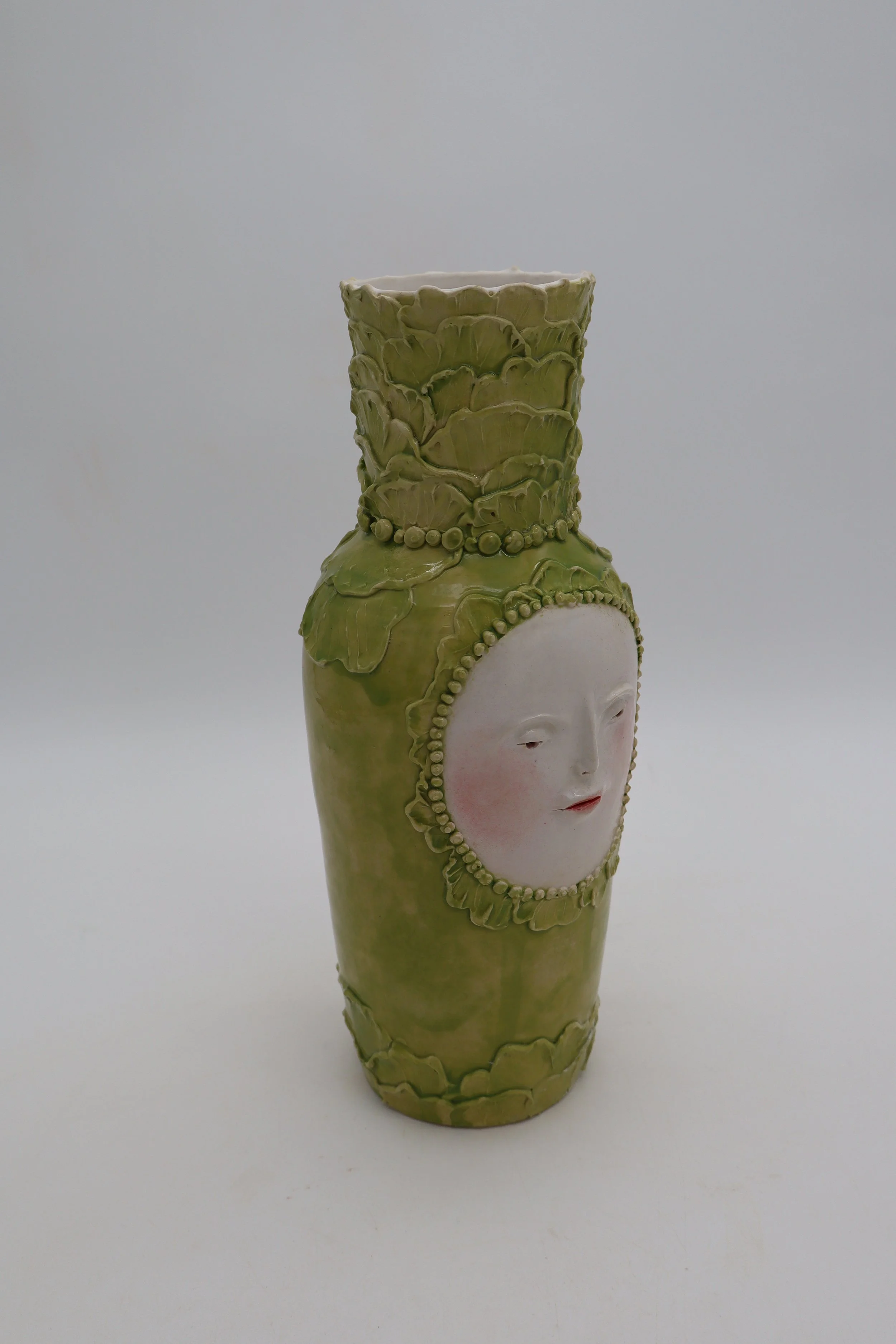 Ceramic vase with an abstract face design on the front, decorated with textured green and white elements, set against a plain background.