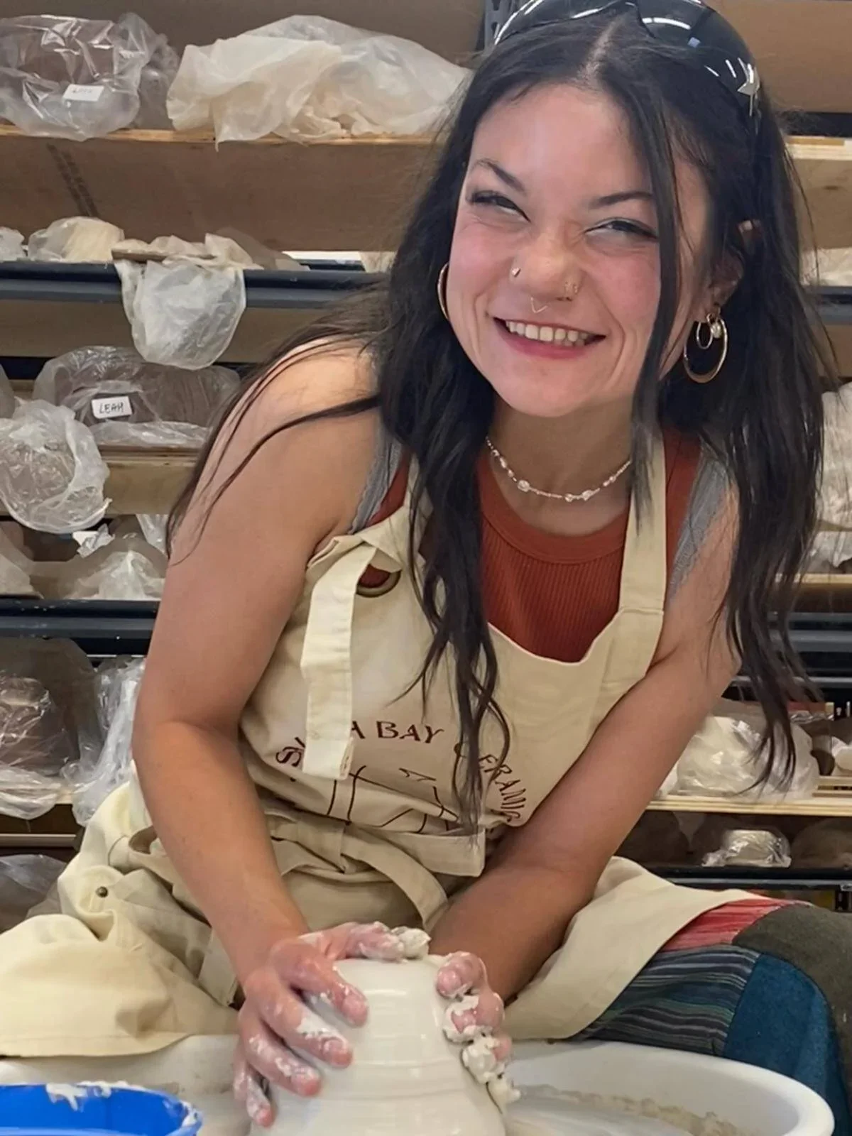 A woman with dark hair, smiling and wearing a beige apron, working on pottery in a studio.