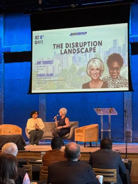 Two women on stage at a conference, one with light hair and the other with dark hair, speaking and facing the audience. A large screen behind them displays the event title 'The Disruption Landscape' with images of two women and details about the event.