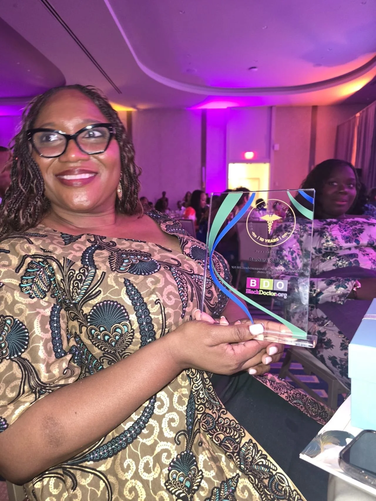 A woman with glasses and curly hair smiling at an event, holding an award plaque with the logos 'B D O' and 'BlackDoctors.org'. She is wearing a patterned dress, and there are other people and purple lighting in the background.