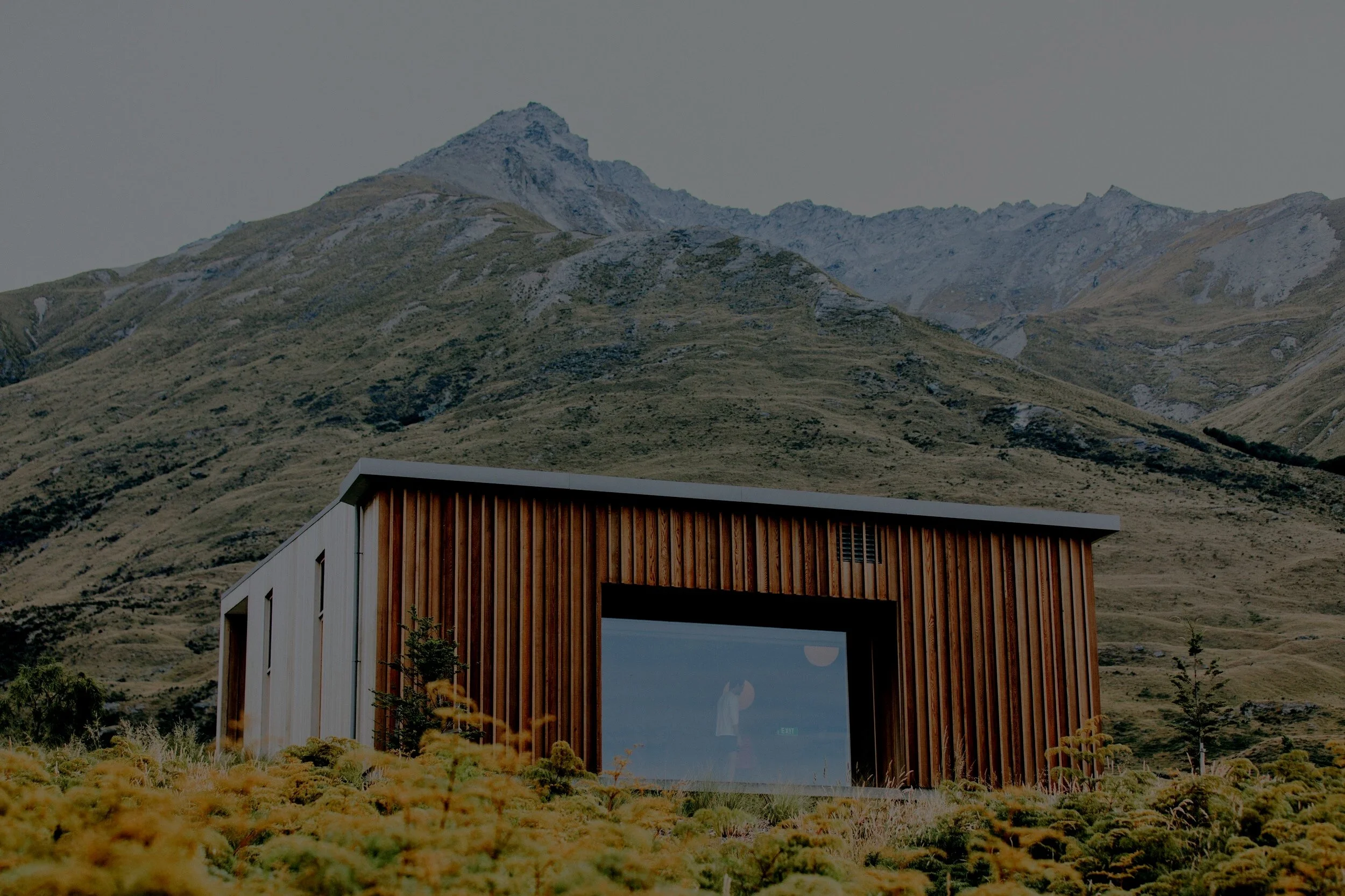 Modern house with large glass window and wooden exterior, situated in a mountain landscape with grassy foreground and snow-capped peaks in the background.
