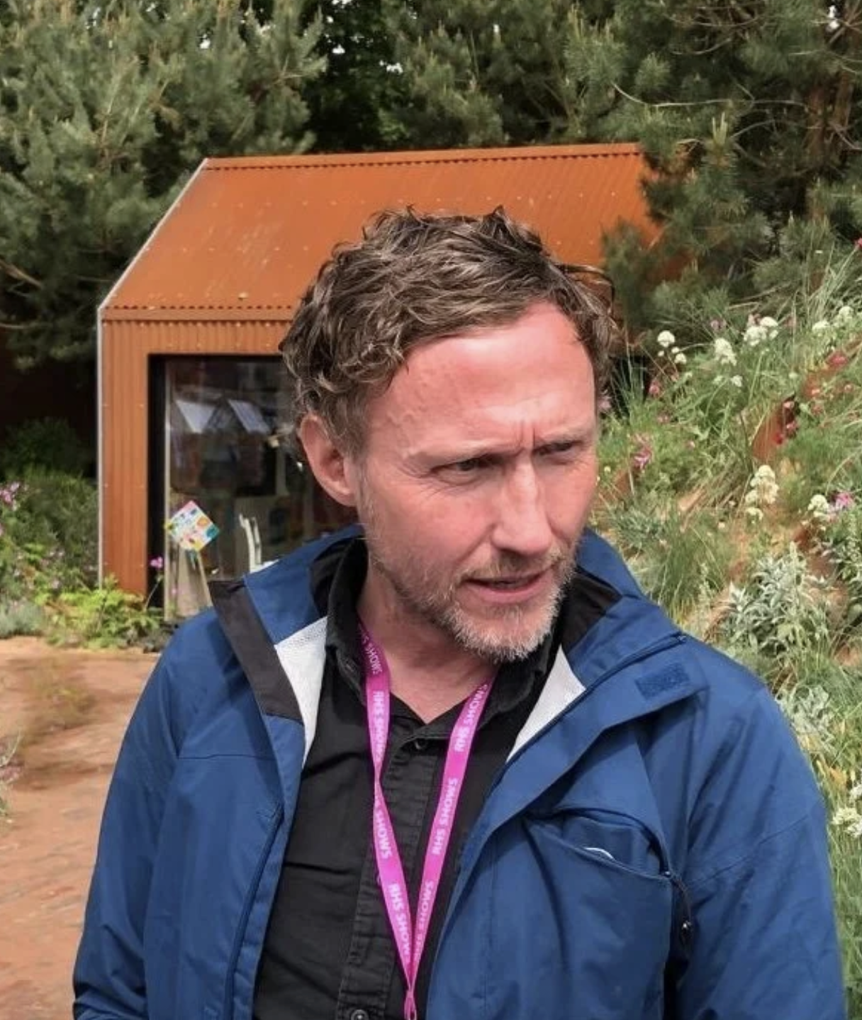 A man with curly hair and a beard, wearing a blue jacket and a black shirt, standing outside near a garden with bushes and flowers, with a small red building or shed in the background.