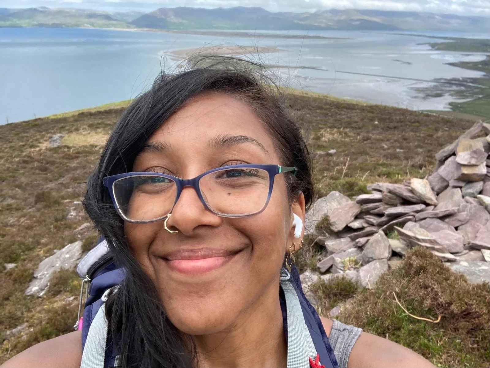 A smiling woman with glasses, a nose ring, and earrings taking a selfie outdoors on a mountain trail, with a view of a lake and hills in the background.