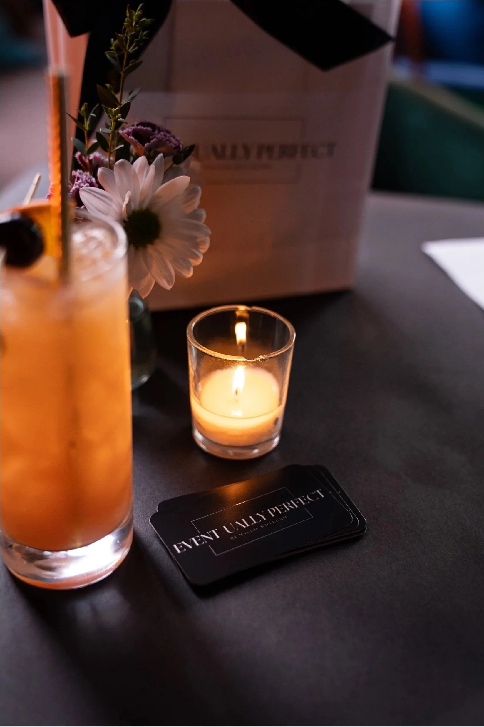 A table setting with a lit candle in a small glass holder, a tall drink with an orange garnish and a flower decoration, a stack of black cards that reads "Event Usually Perfect," and a partial view of a cardboard box in the background.