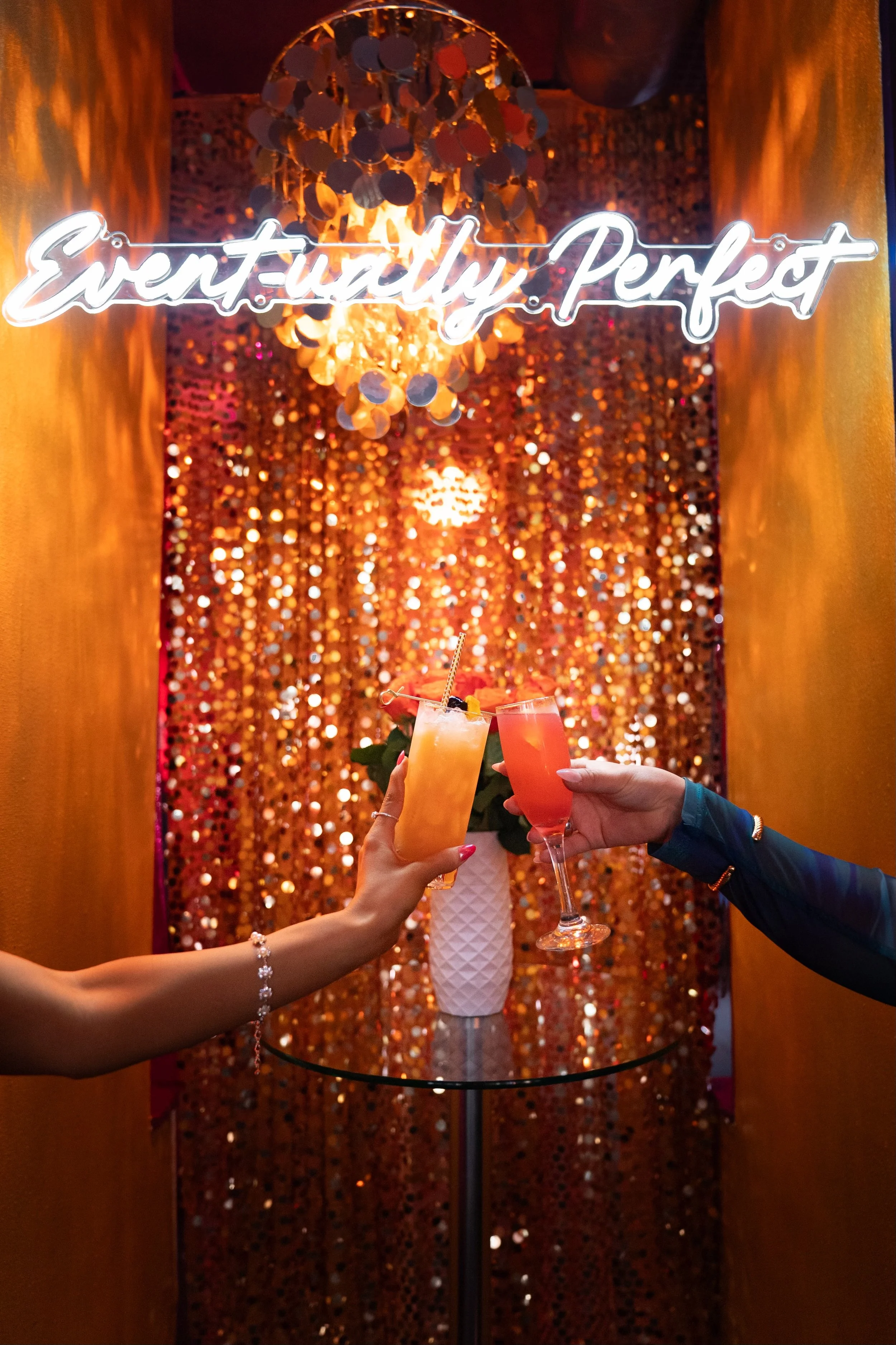Two people holding colorful cocktails as they toast in front of a decorative, sparkly background with a chandelier and a neon sign that reads 'Eventually Perfect'.