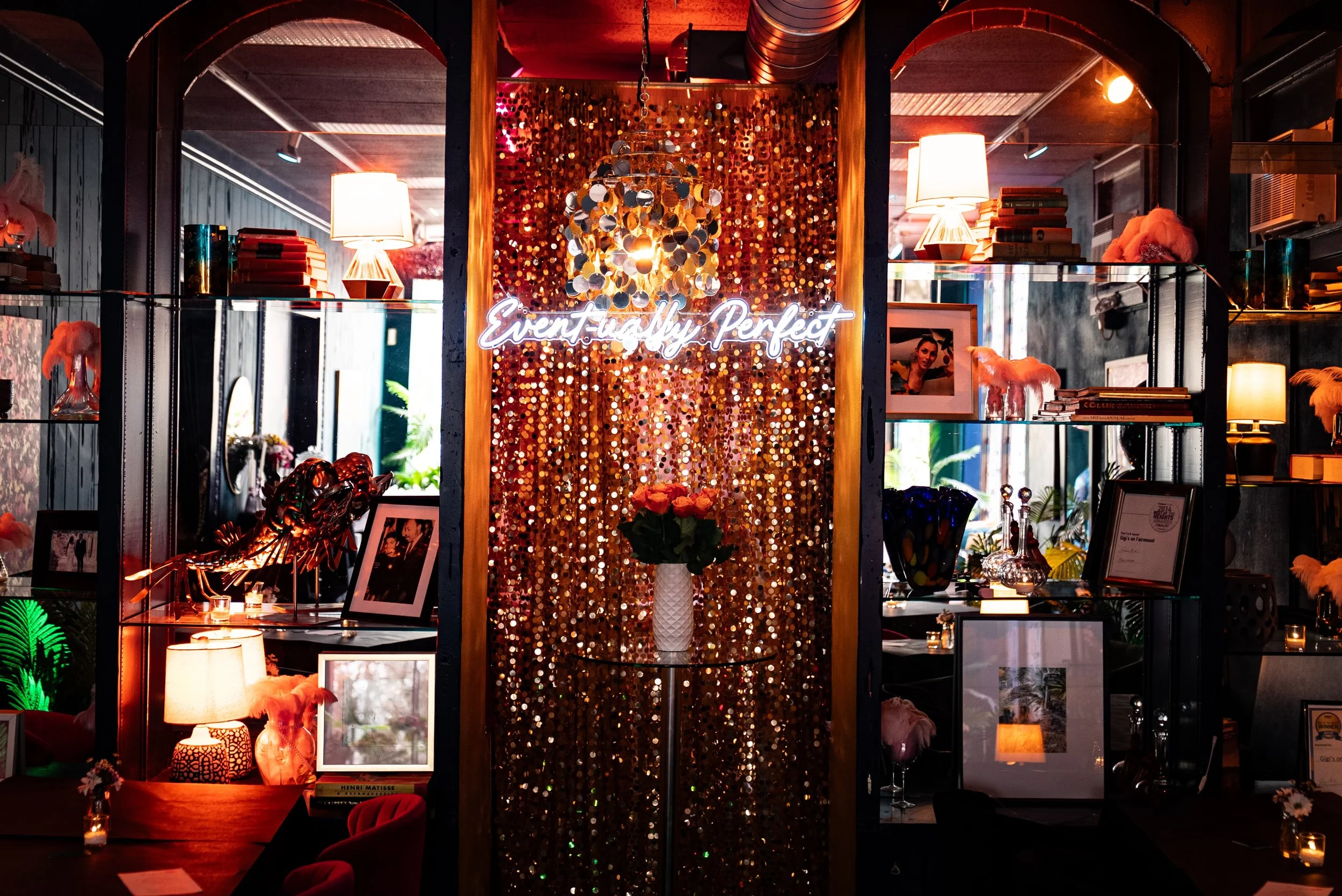 A decorated interior space with warm lighting, shelves holding books and decorative objects, a neon sign saying 'Eventfully Perfect,' and a sequin curtain background.