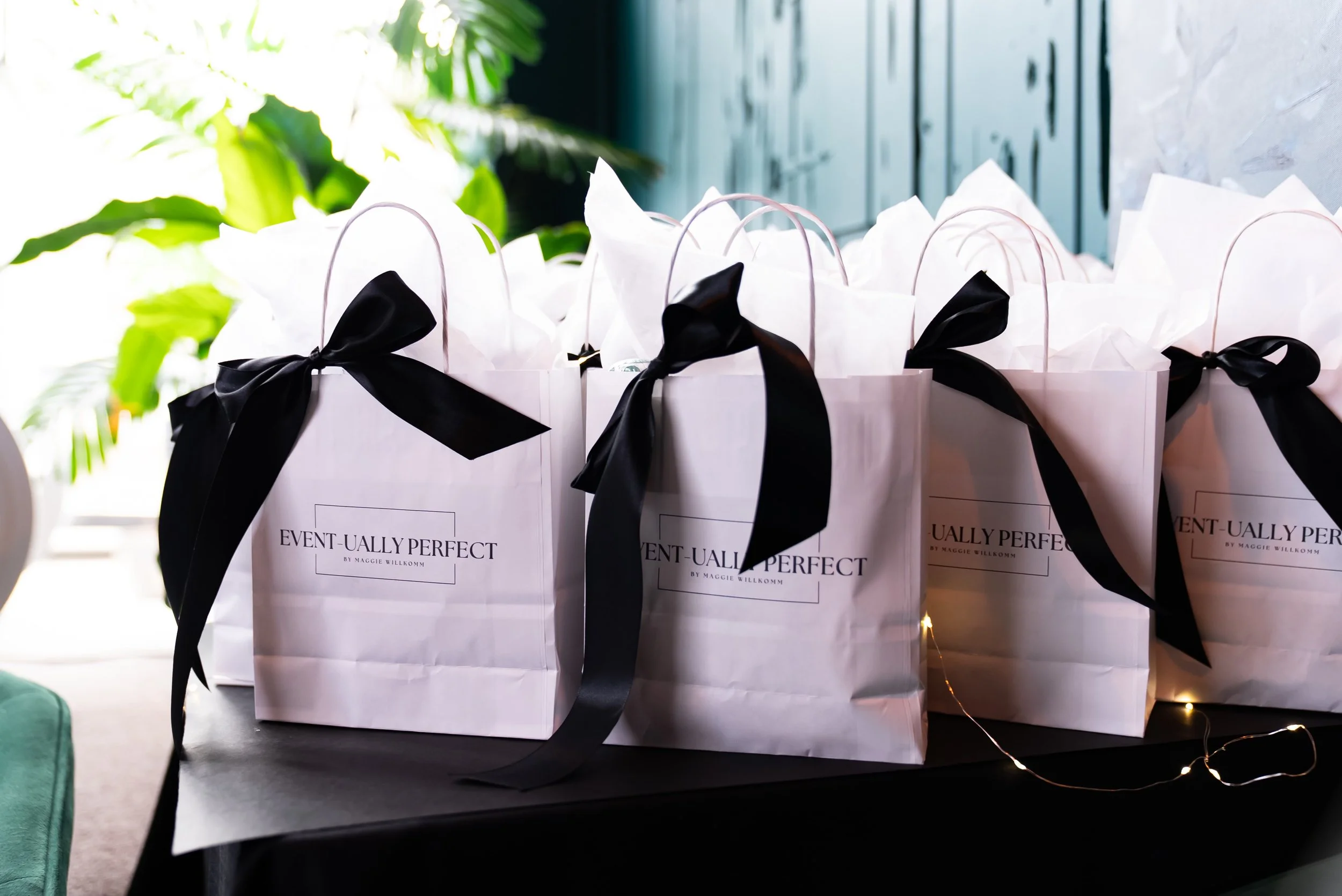 White gift bags with black ribbons on a table, printed with 'Event-Ually Perfect by Maggie Wilkom'.