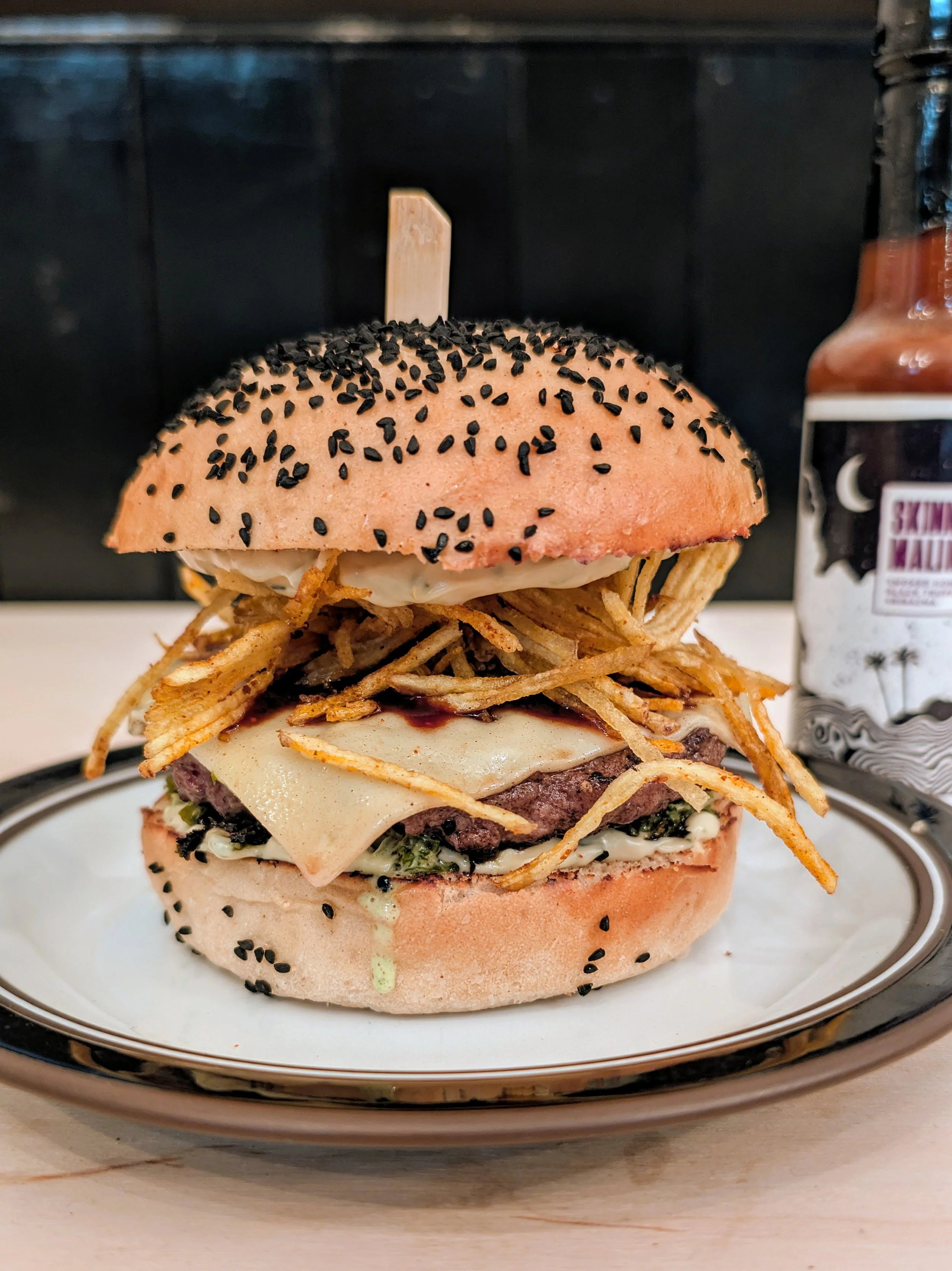 A large burger with a seed-topped bun, crispy noodles, melted cheese, a beef patty, and vegetables, placed on a black and white plate.