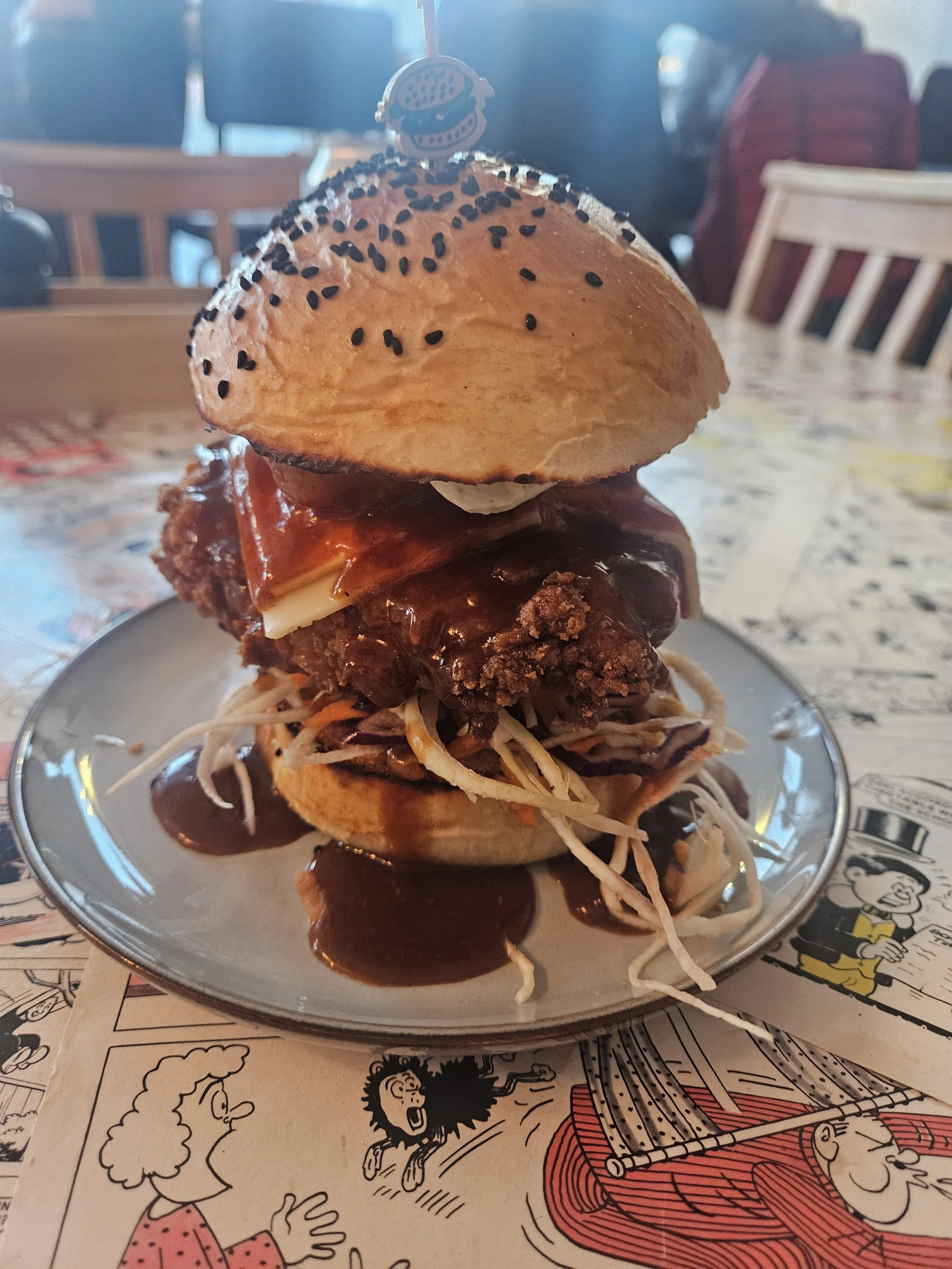 A chicken burger, with basil miso mayo, house slaw, KBBQ sauce, and crispy noodles, served on a plate, placed on a comic strip-themed table.