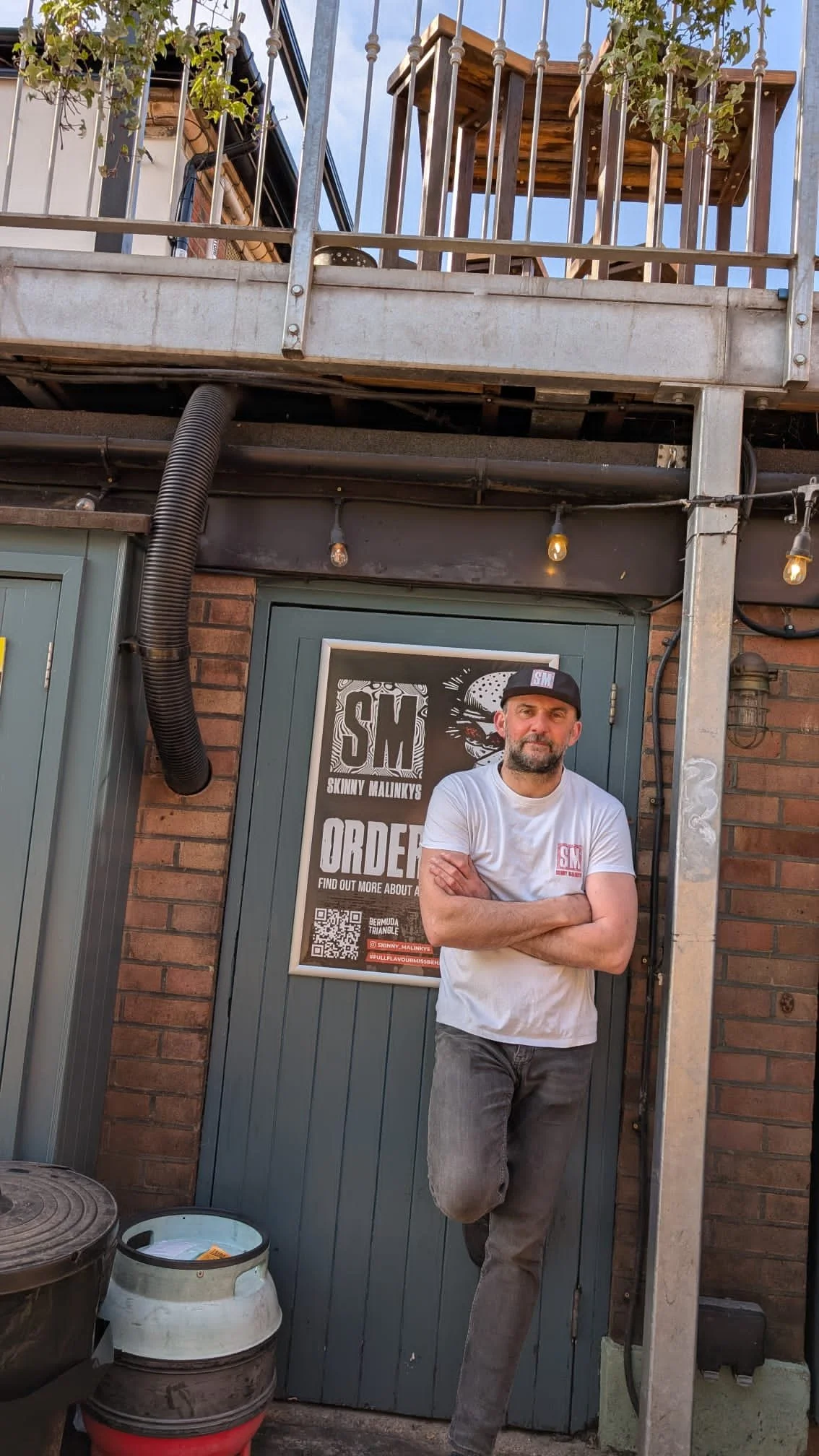 A man with crossed arms leaning against a door outside a building with a poster on it. The poster has the text 'SKINNY MALINKYS ORDER' and a QR code. The man is wearing a white T-shirt, gray pants, and a black cap. There are outdoor lights hanging above and some barrels and containers on the ground in front, with a brick wall and metal railing above.