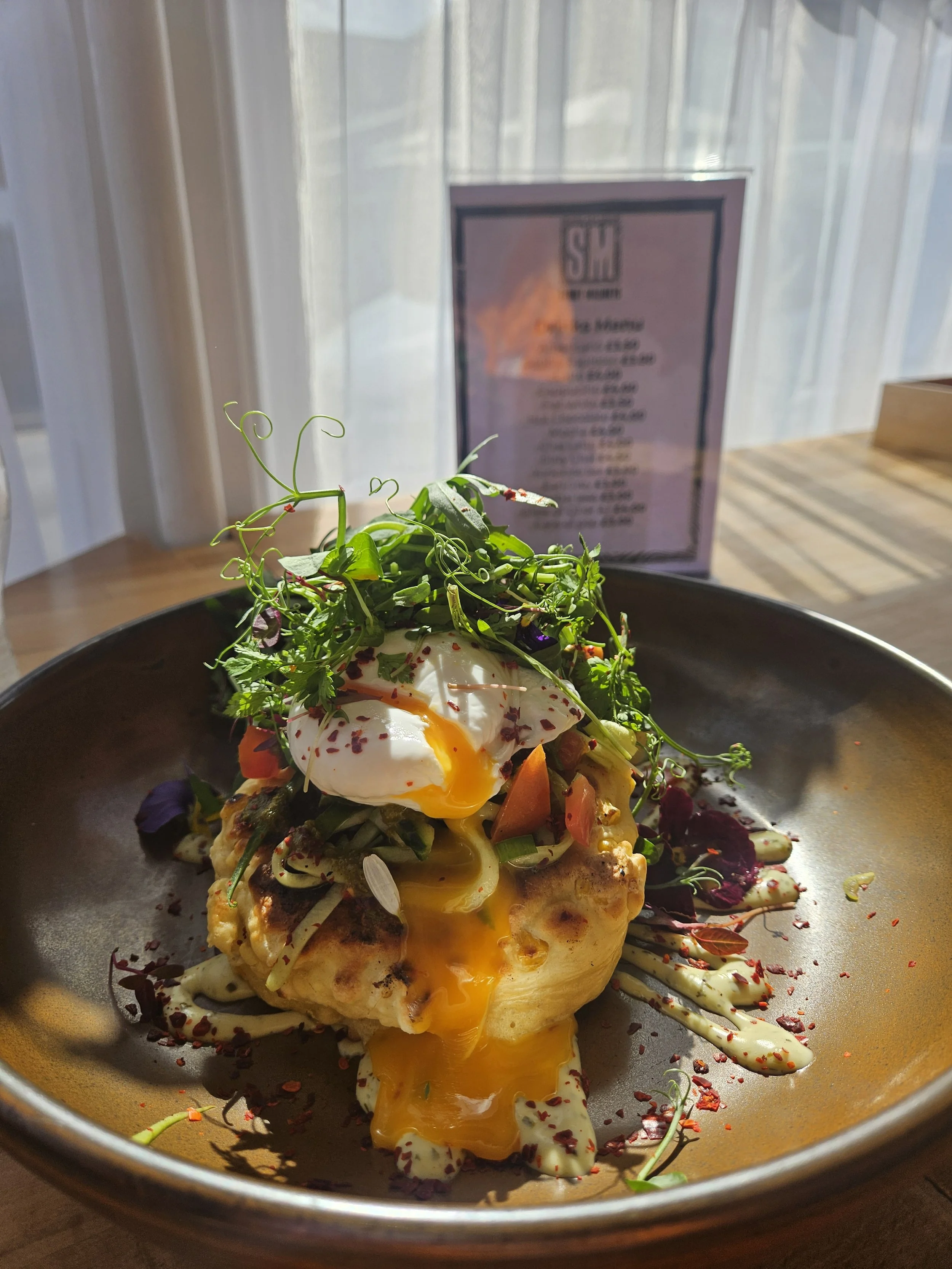 A savory breakfast dish featuring a baked egg on top of vegetables, garnished with microgreens, served on a dark plate with a condiment sauce.