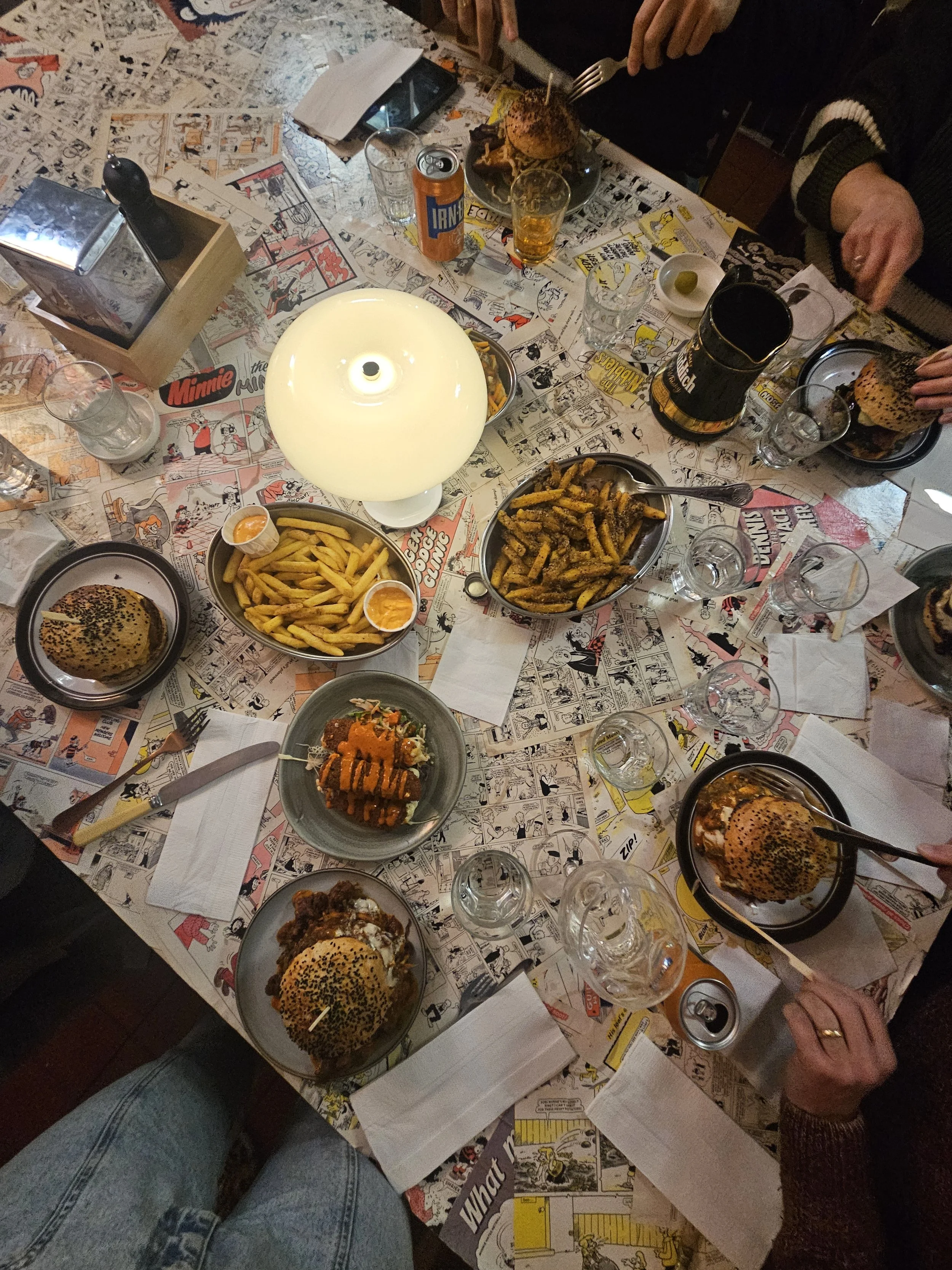 A table with comic strip newspaper pages, holding burgers, fries, and drinks, surrounded by people eating.
