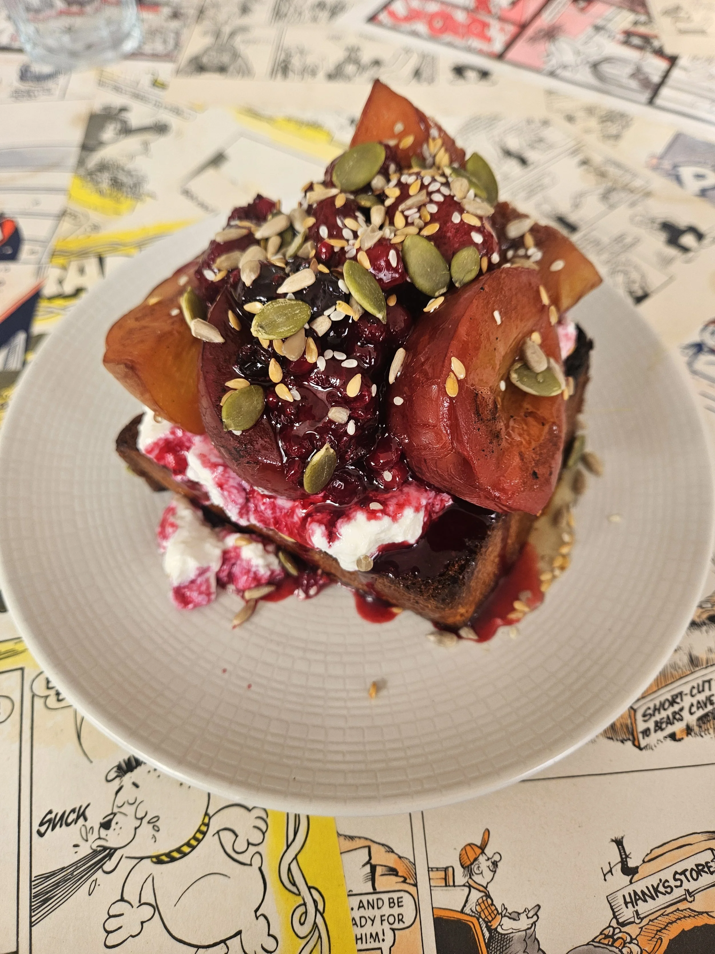 Honey butter toast topped with mixed berries, seeds, and ricotta, on a white dish, placed on a table decorated with comic strip pages.