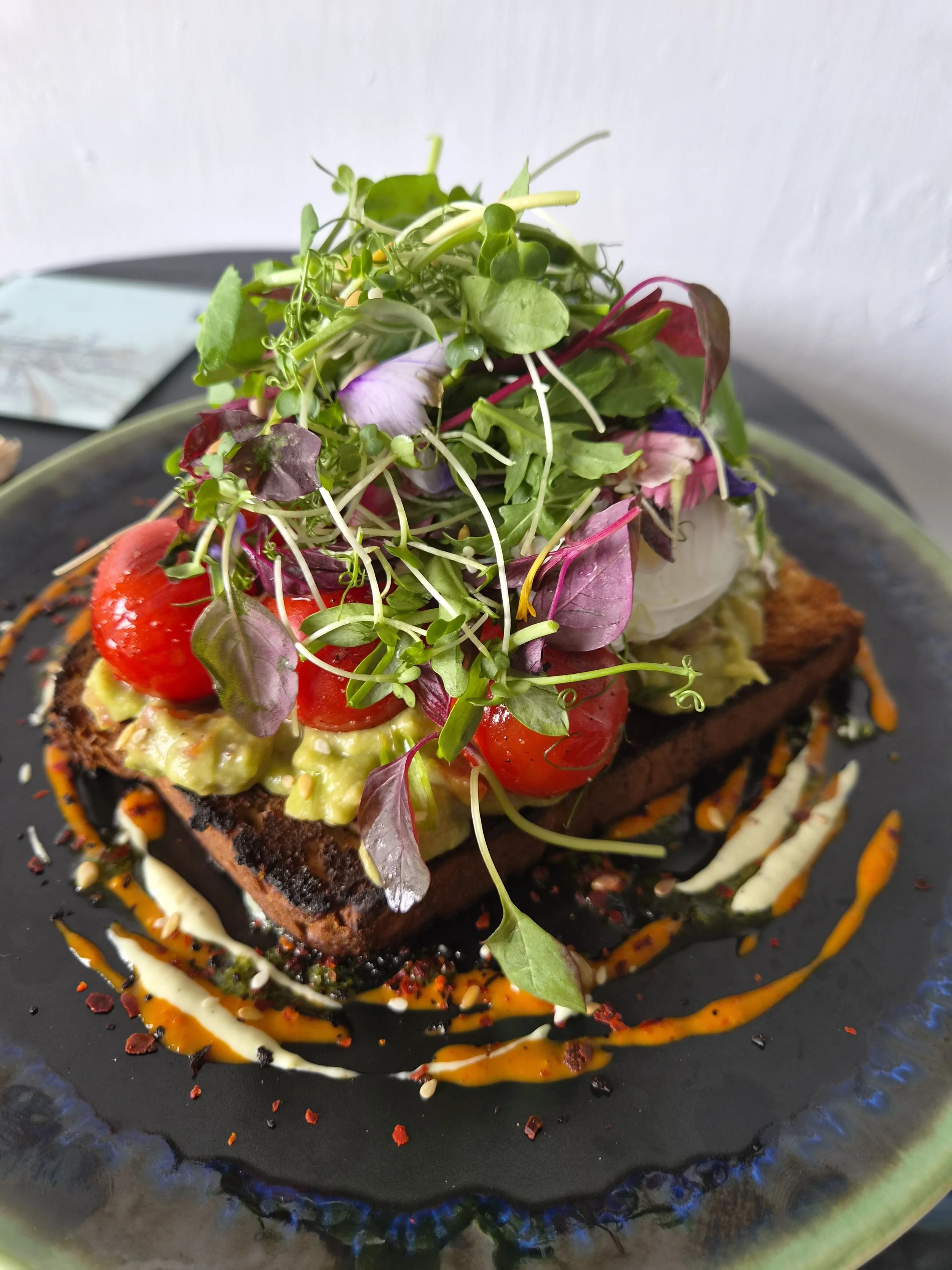 A dish with a slice of toast topped with mashed avocado, cherry tomatoes, microgreens, and garnished with drizzles of sauces on a black plate.
