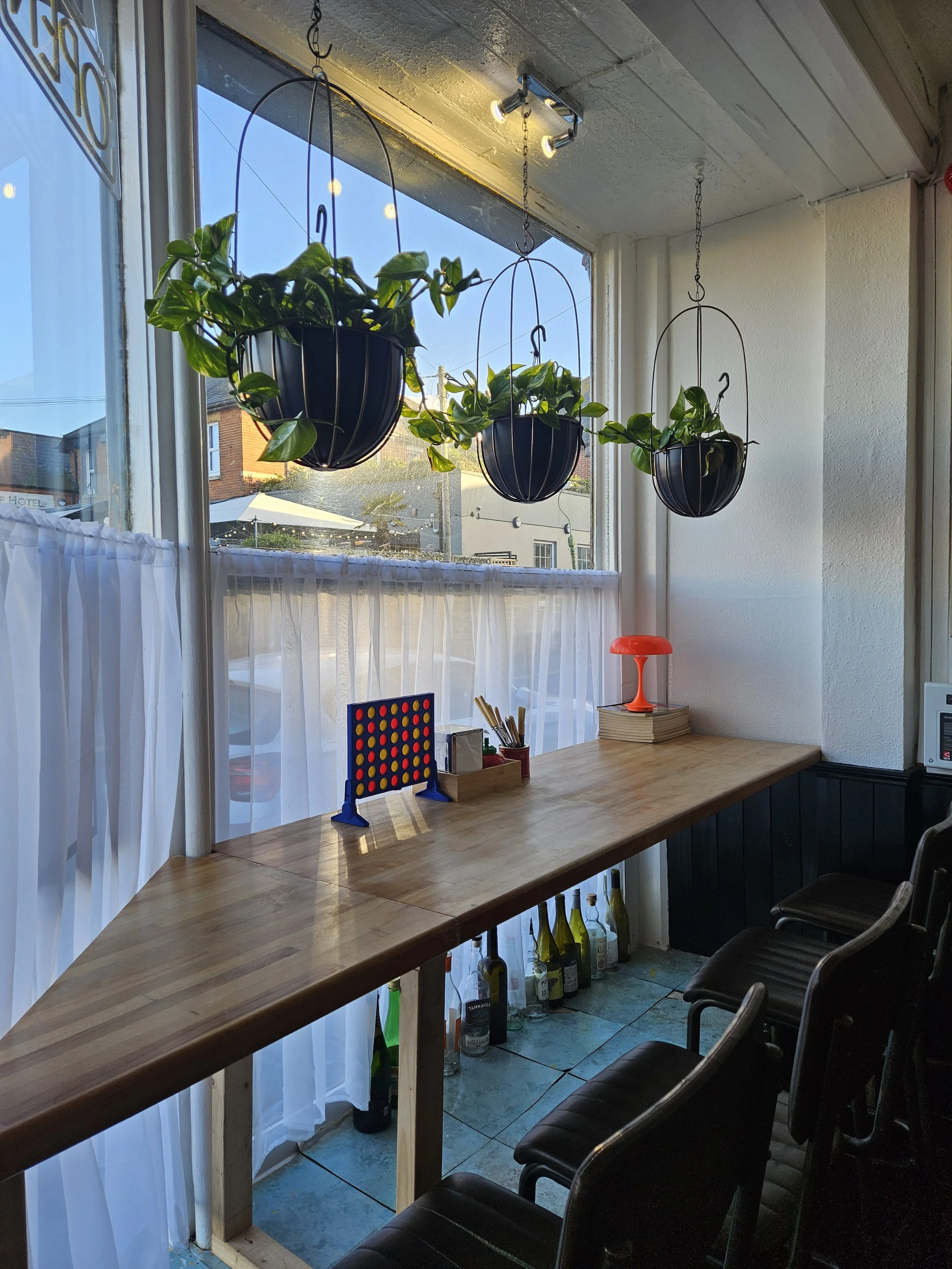 A cosy indoor dining area with a wooden table, black chairs, hanging plant baskets with green plants, a small game board on the table, with large windows letting in natural light.