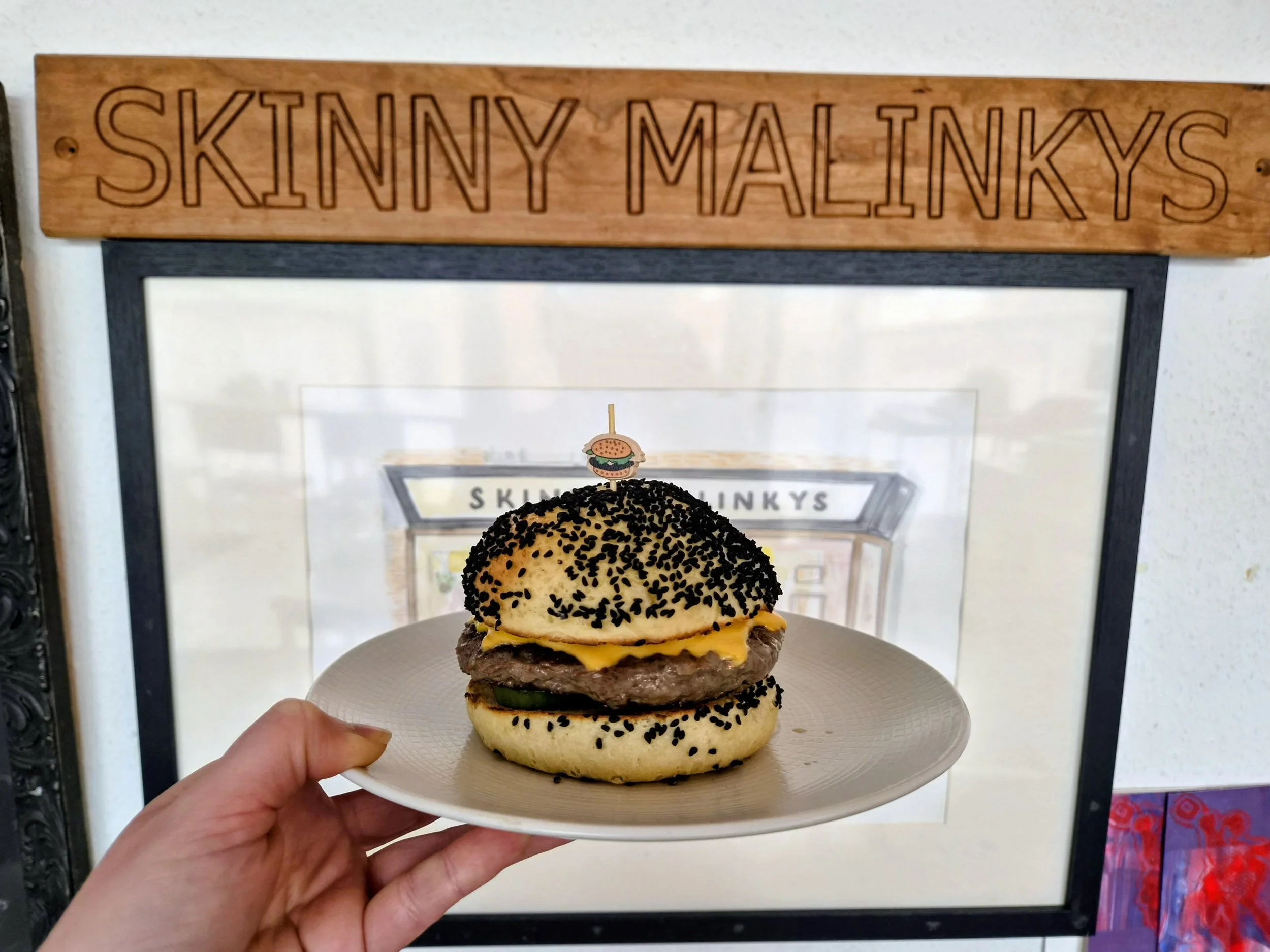 The OG - a cheeseburger with American style cheese, a beef patty, pickles, onion, ketchup, fermented chill mustard, in a potato bun, being held on a white plate, with a sign that reads 'SKINNY MALINKYS' in the background.