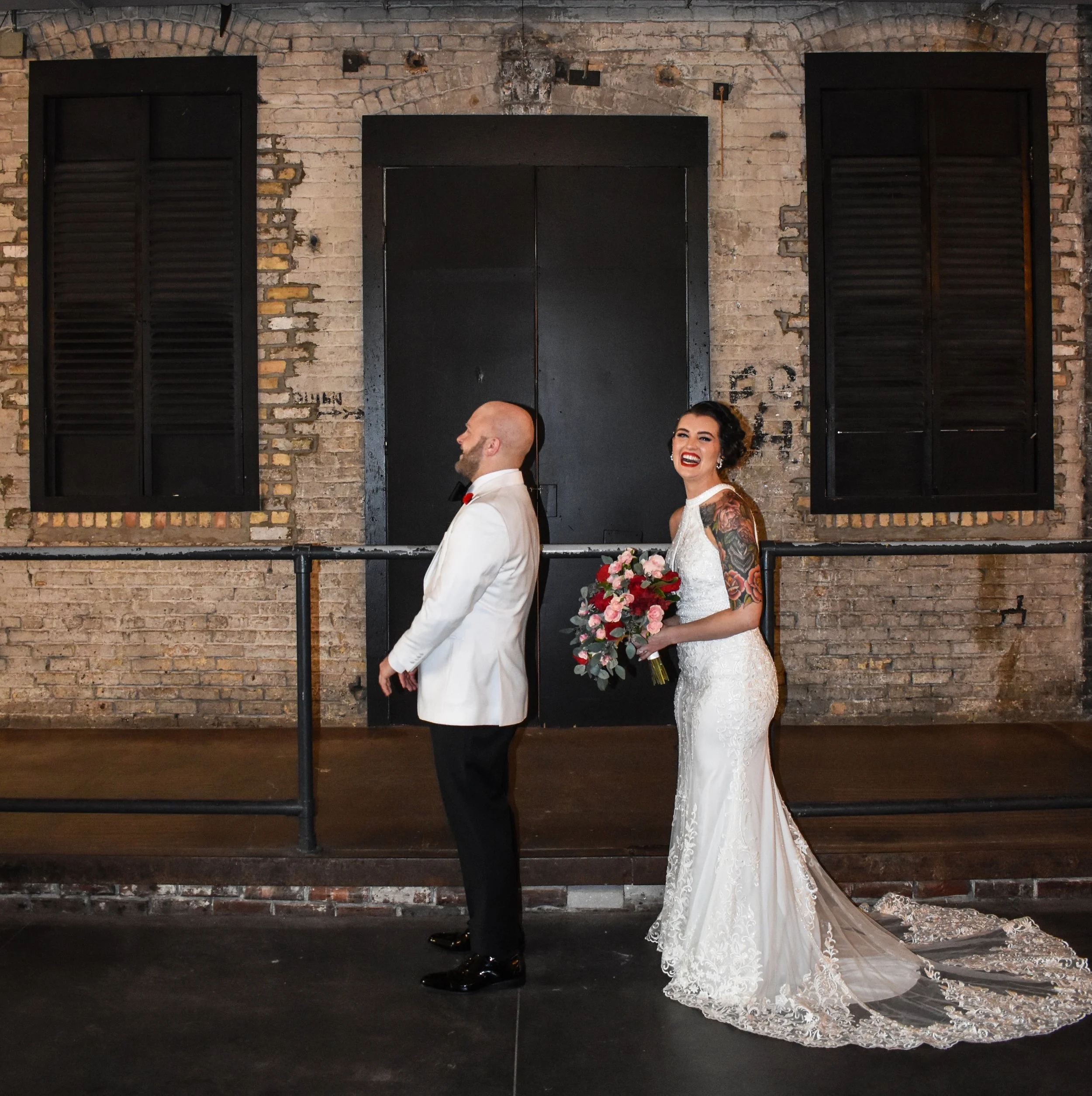 A bride in a lace wedding dress with tattoos on her arms holding a bouquet of pink and red flowers, smiling, standing behind a groom in a white tuxedo with a red boutonniere, both facing opposite directions in front of an industrial brick wall with b
