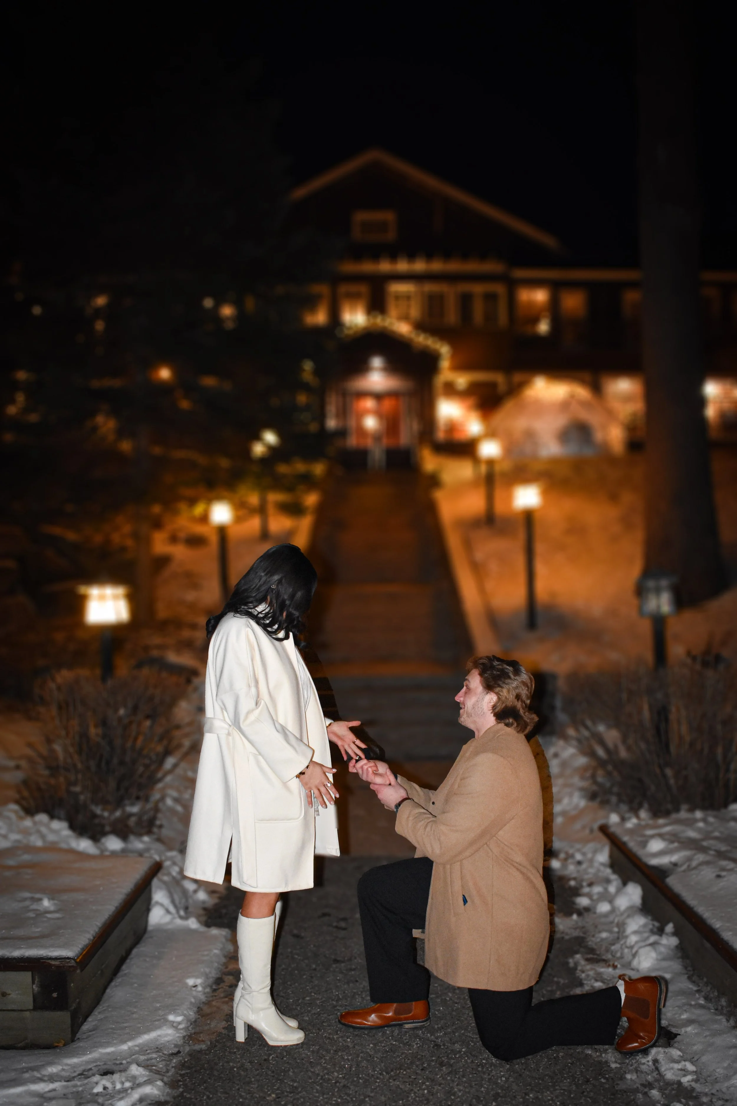 A man proposing to a woman outdoors at night, with a warmly lit house in the background and snow on the ground.