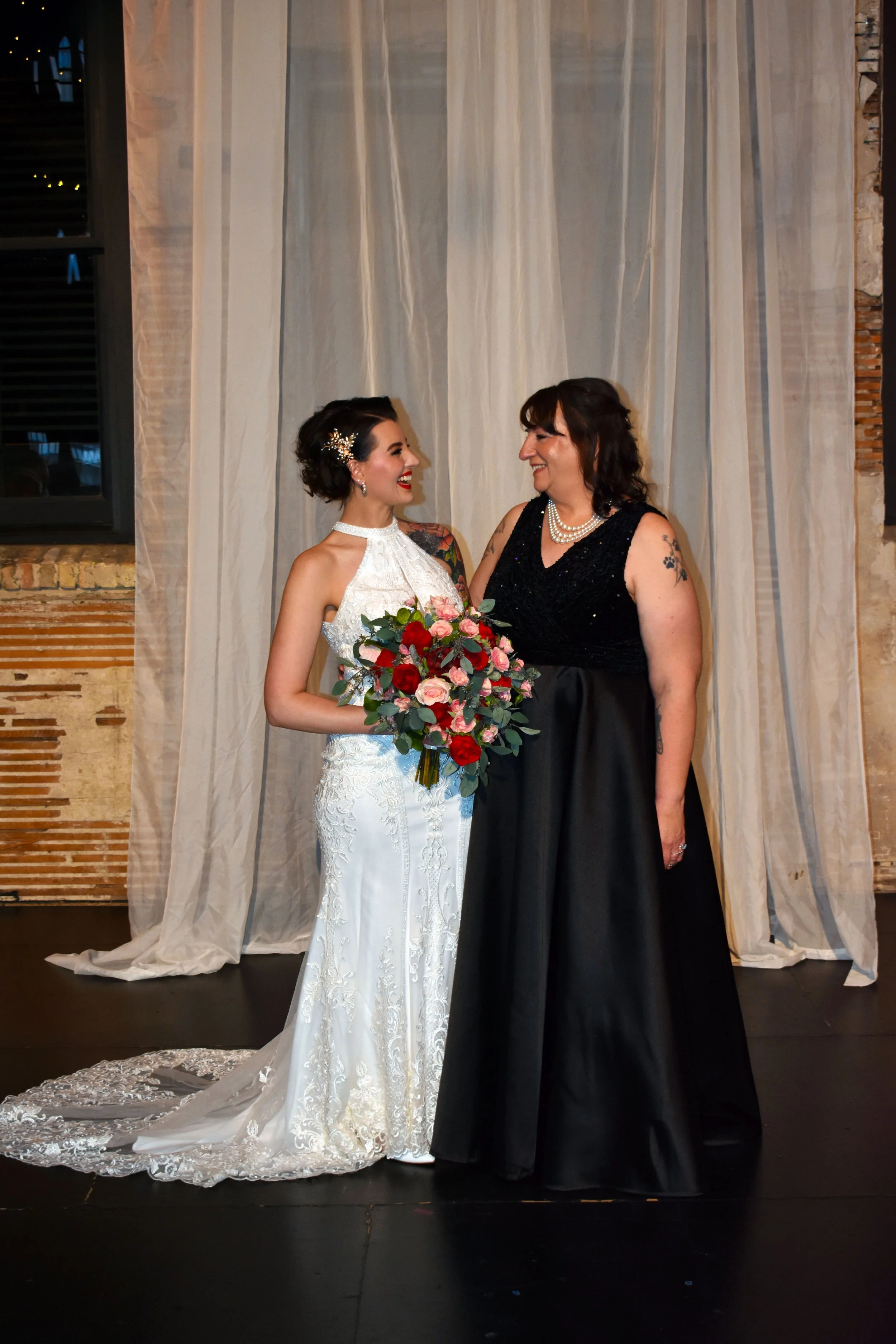 Two women, a bride in a white wedding gown holding a bouquet of pink and red flowers, and a woman in a black dress, smiling at each other against a beige curtain backdrop.