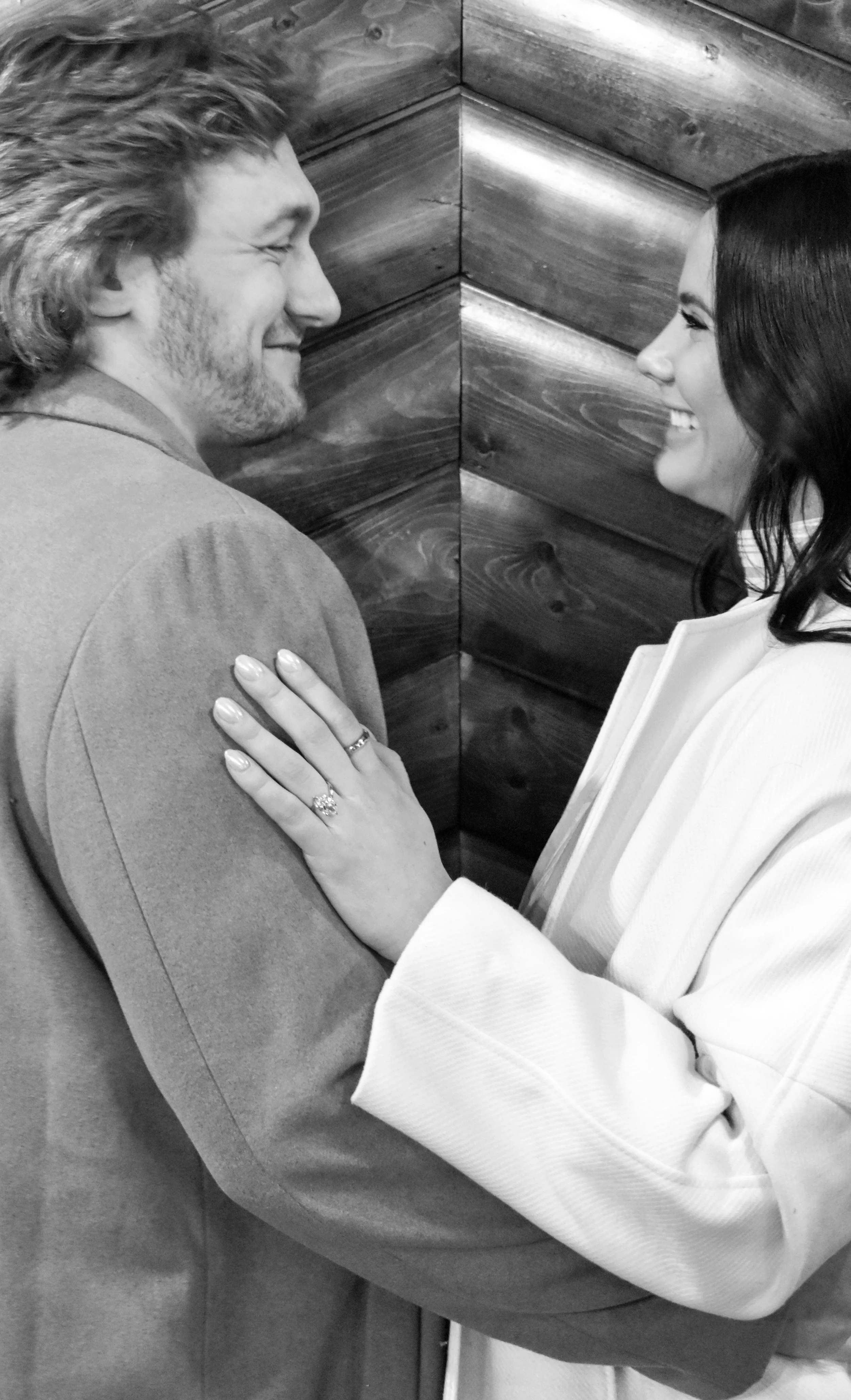A couple smiling and looking at each other, with the woman touching the man's shoulder. The woman is wearing a white jacket and has a ring on her finger.