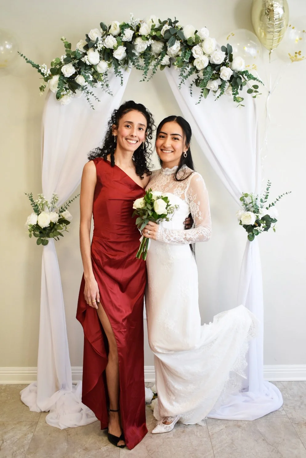 Two women, one in a white wedding dress holding a bouquet and the other in a red gown, smiling and standing in front of a floral arch at a wedding celebration.