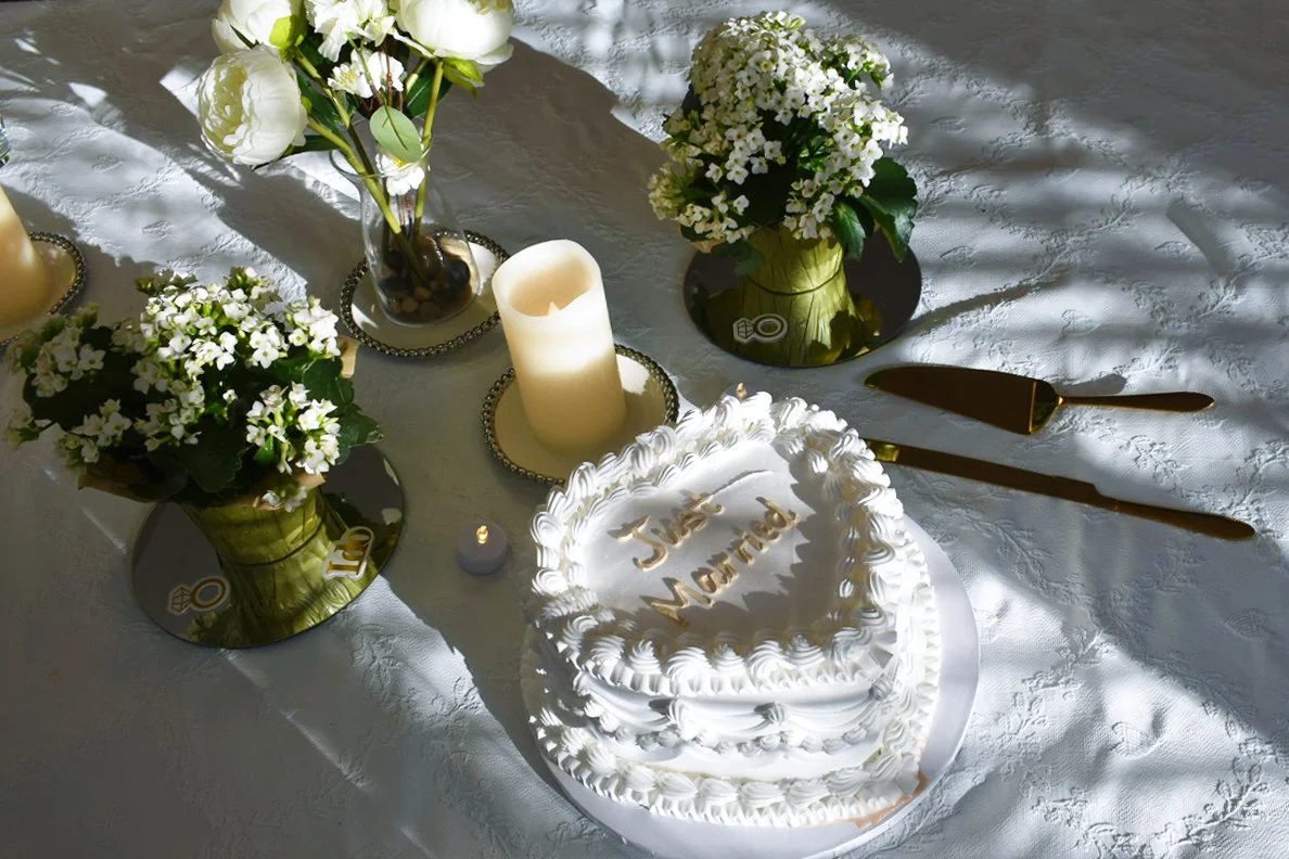 Heart-shaped wedding cake with white icing and gold lettering that says "Just Married" surrounded by white flowers, candles, and golden utensils on a white textured tablecloth.