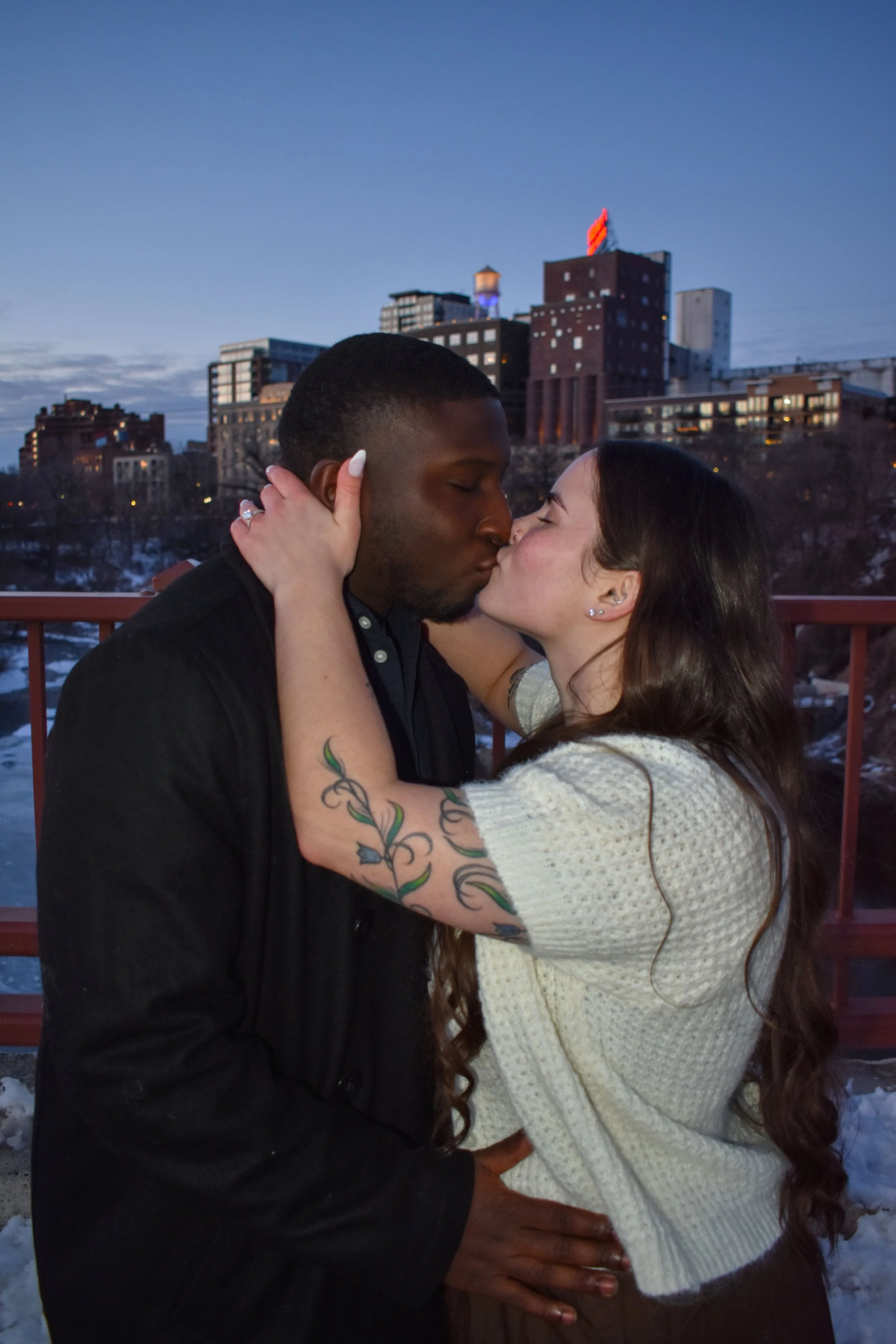 A couple sharing a kiss on a bridge at sunset with city buildings in the background.