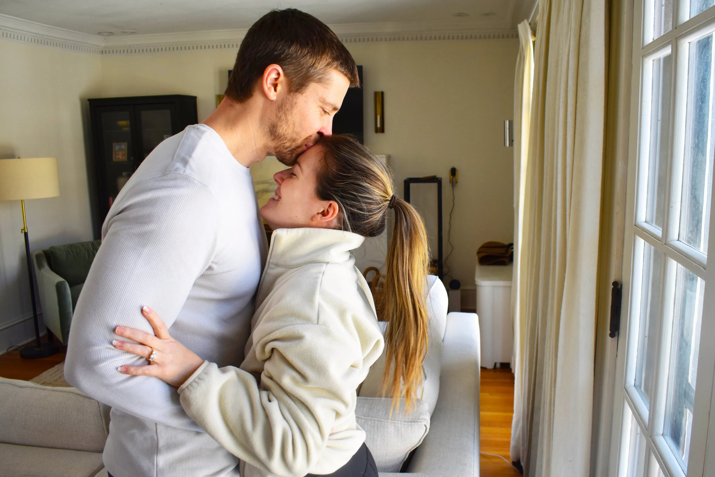 A couple sharing an intimate embrace in a living room, with large windows and natural light.
