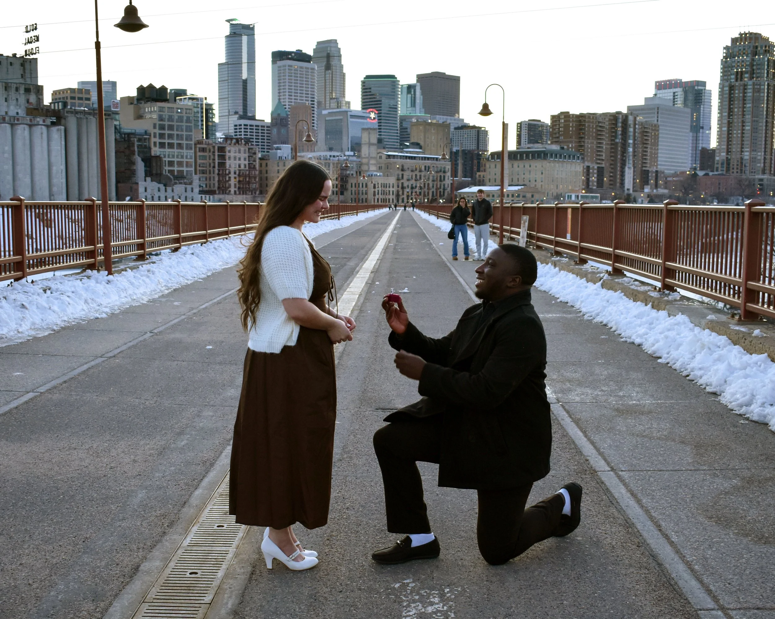 A man proposing marriage to a woman on a snowy city bridge, with the man kneeling and offering a ring in a red box, while the woman smiles and looks at him.