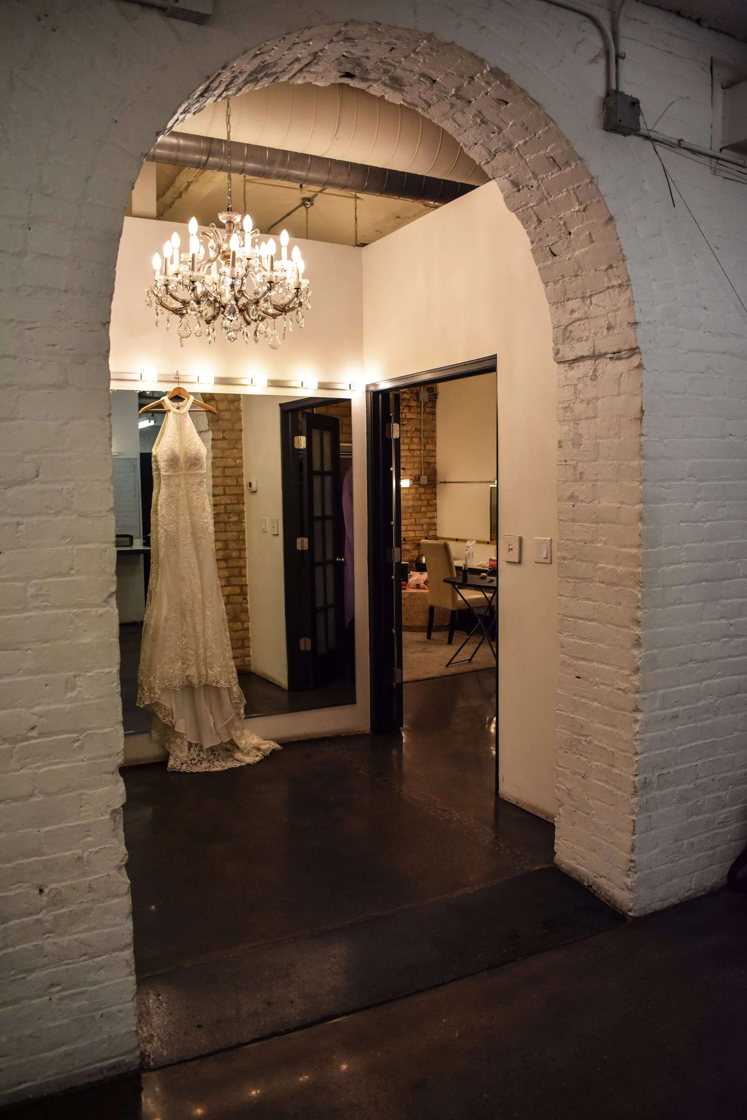 An arched brick doorway opening into a room with a white wedding dress hanging, a chandelier, and a modern interior with exposed brick walls.